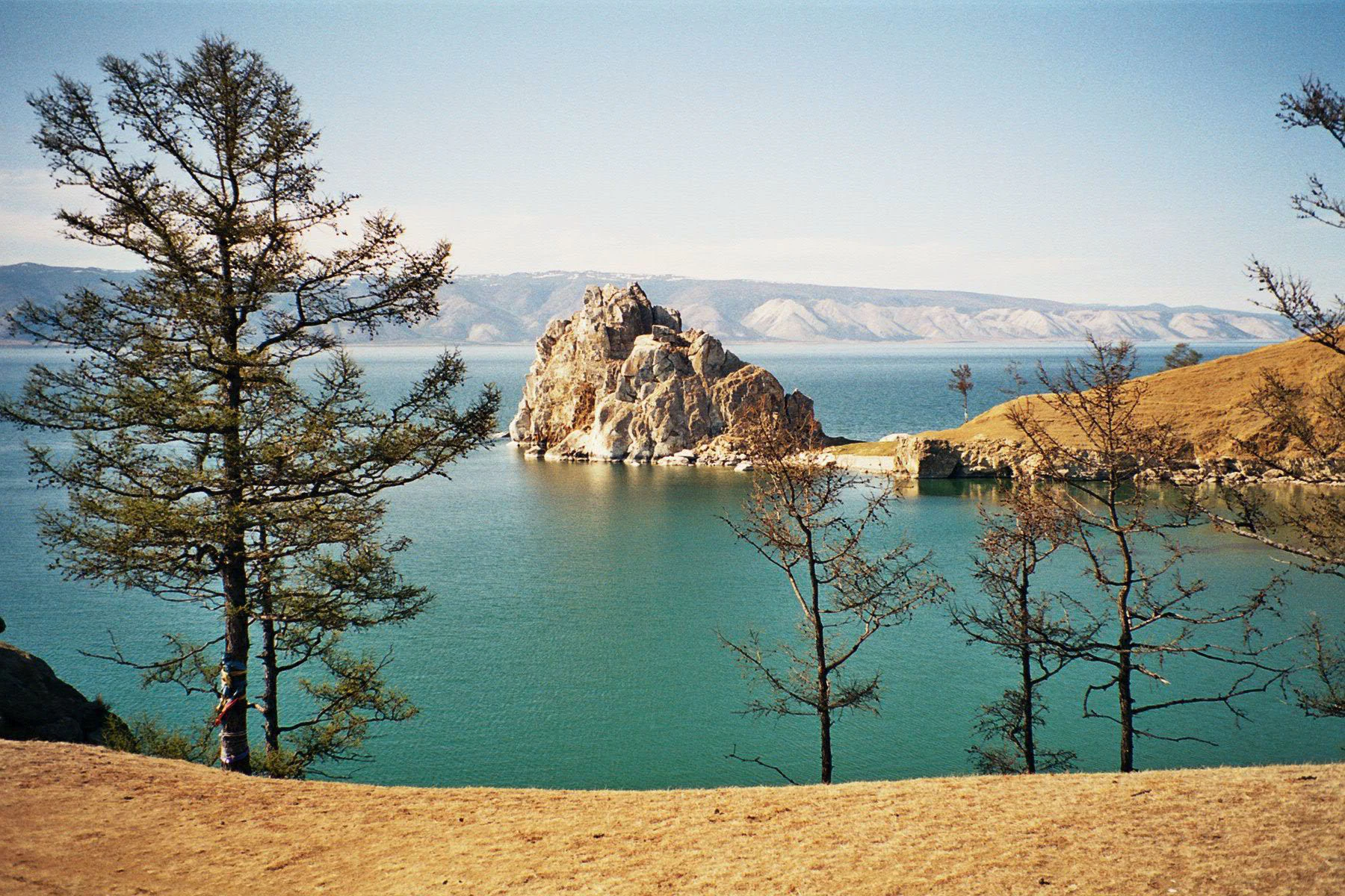 A large, craggy limestone rock formation stands in the blue water of a lake, with dry grassy hills and scattered thin trees in the foreground and a hazy mountain range in the distant background.