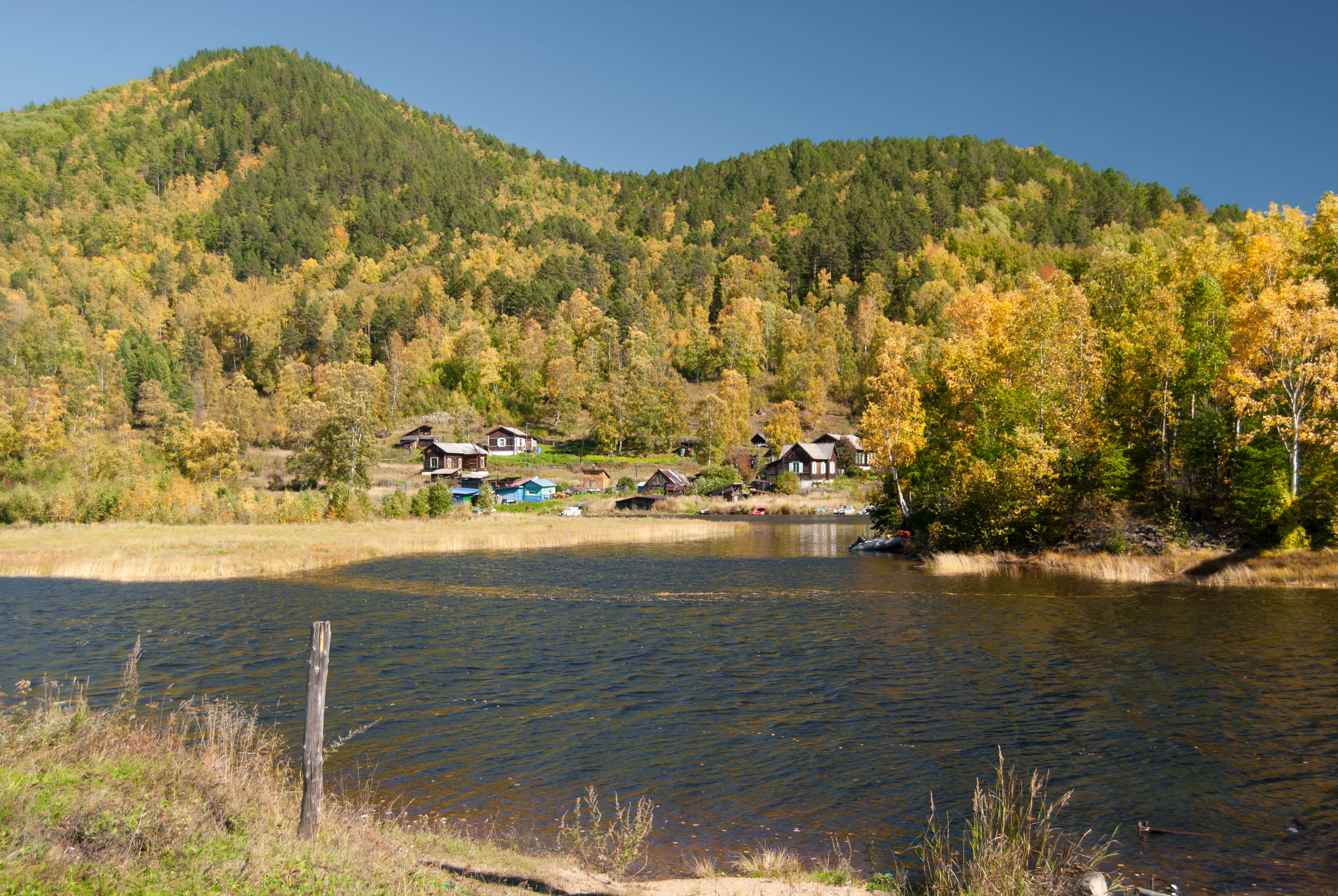 A wide shot showing a small cluster of rustic wooden buildings nestled at the base of a forested mountain slope, with a calm body of water in the foreground and golden autumn foliage on the trees.