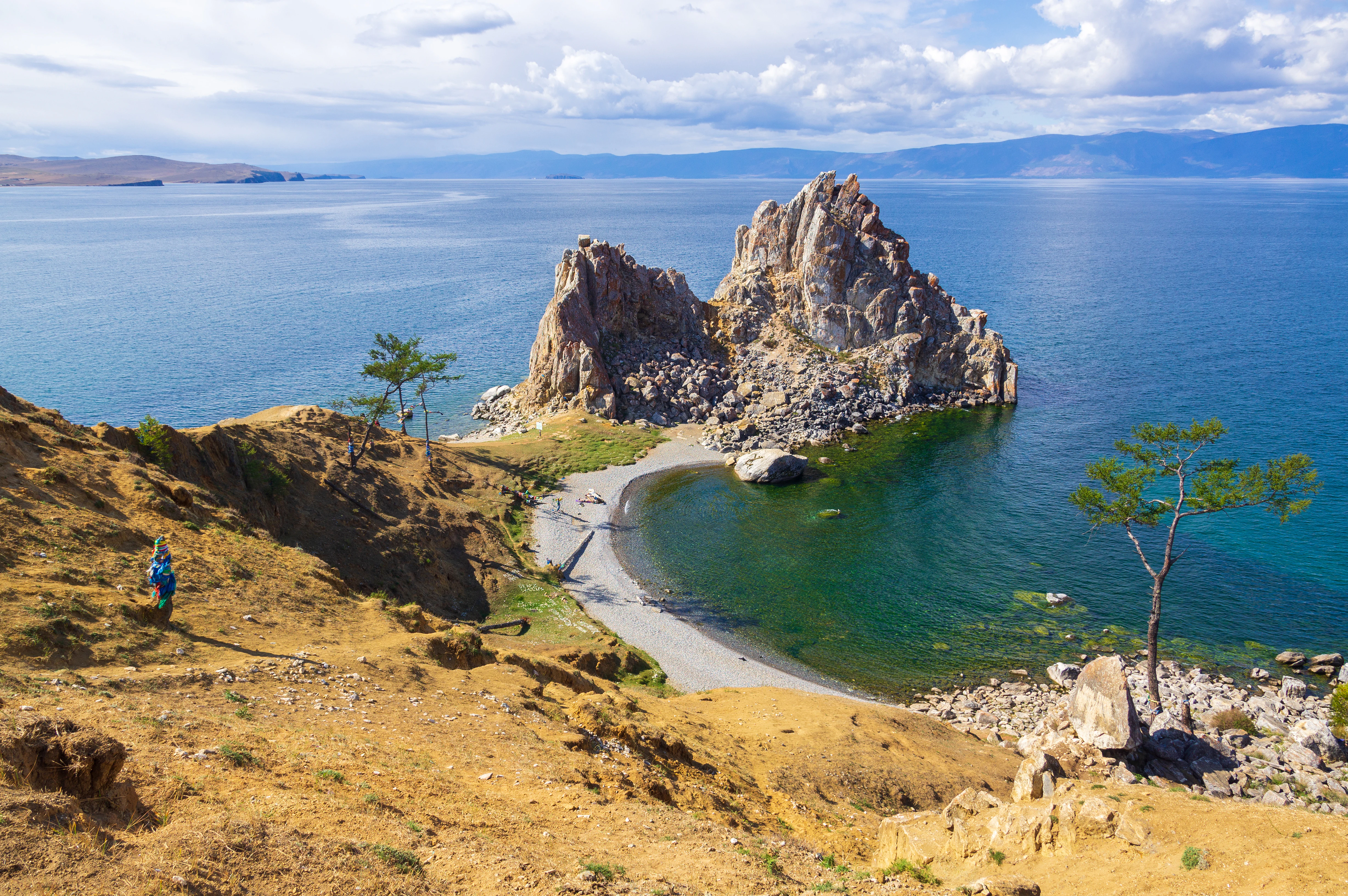 Rocky cape with large stone formation at the edge of Lake Baikal, sandy path along the shore, two trees on a grassy hillside, one person walking