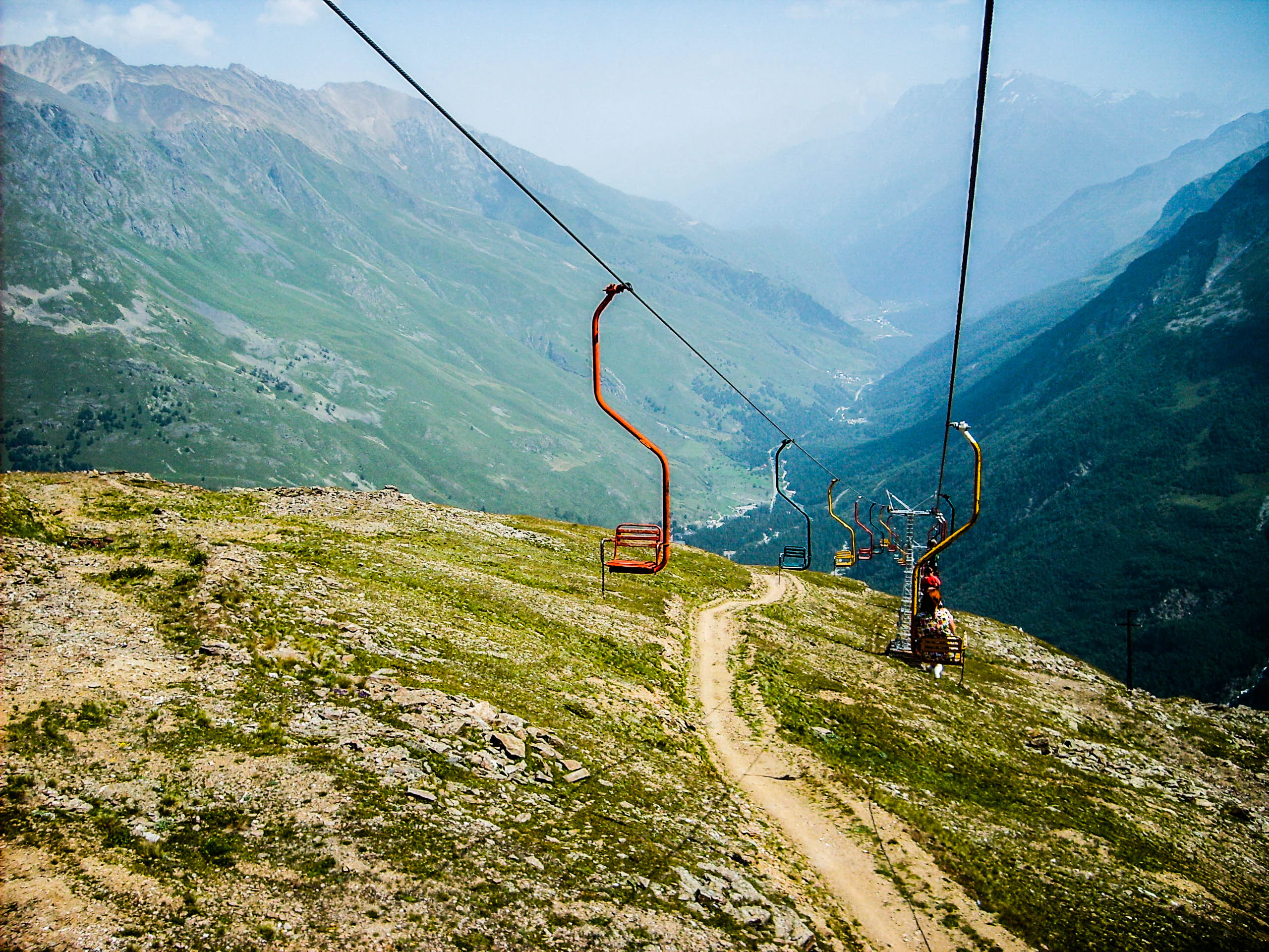 Red cable car chairs suspended by cables over a dirt path, with mountainous landscape and valley in background