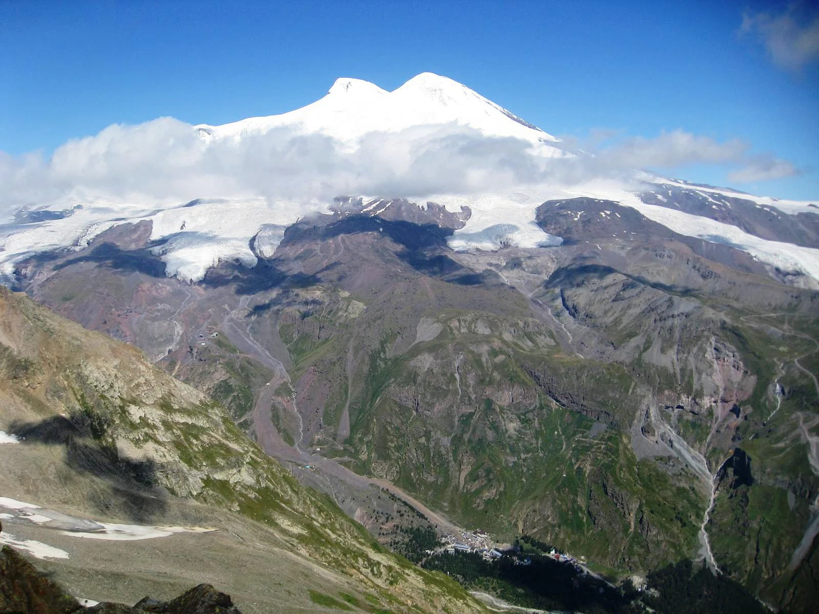 Wide scenic view of Mount Elbrus with snow-capped peaks above a valley with green slopes and rocky terrain