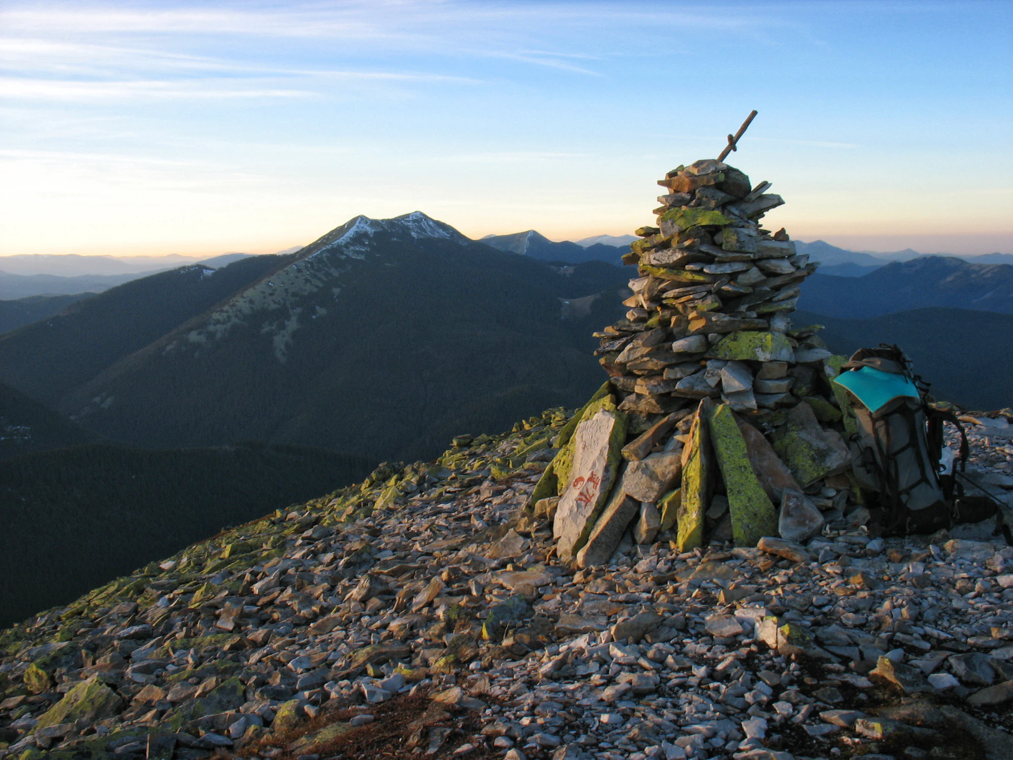 A stacked stone cairn sits on a rocky mountain summit at sunrise, with a hiking backpack nearby and a larger mountain peak in the background.