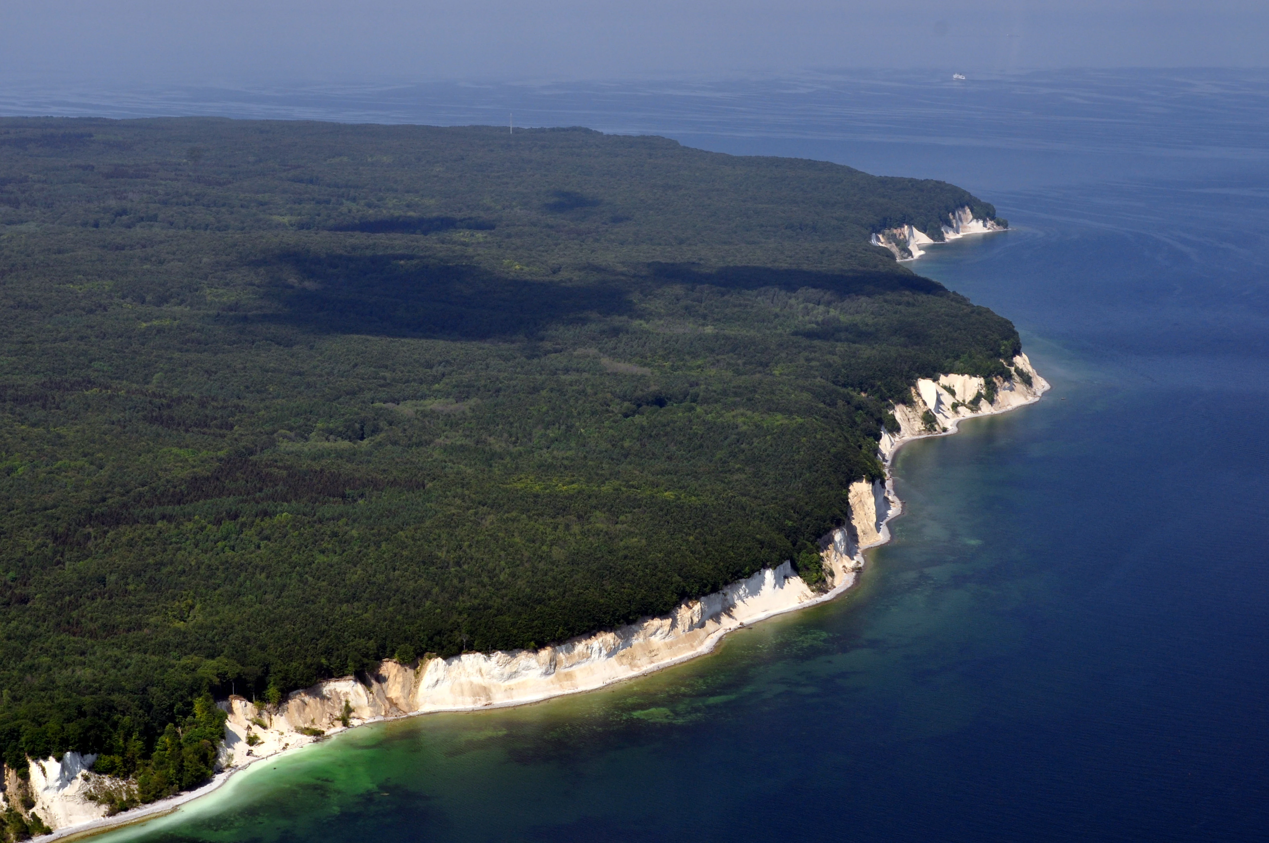 An aerial view showing a steep, white chalk cliff line bordering a dense dark green forest, meeting the deep blue waters of the Baltic Sea.