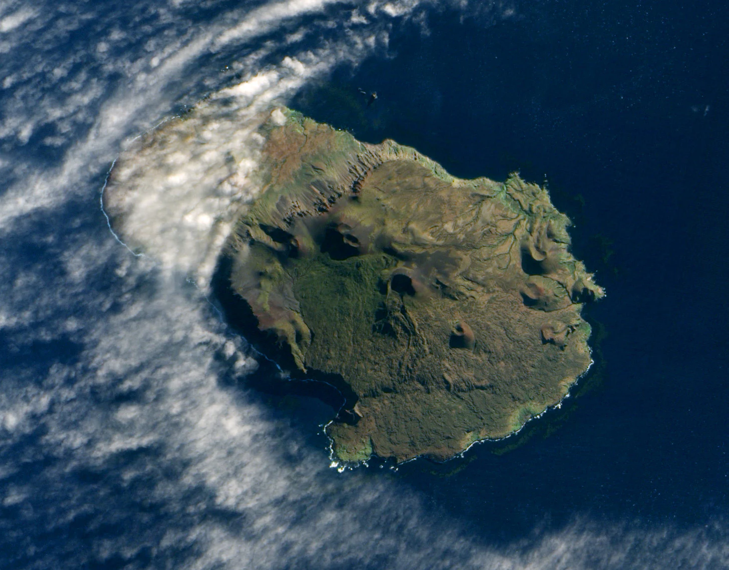 Satellite image from above showing the rugged, volcanic terrain of Prince Edward Island surrounded by ocean and scattered clouds.
