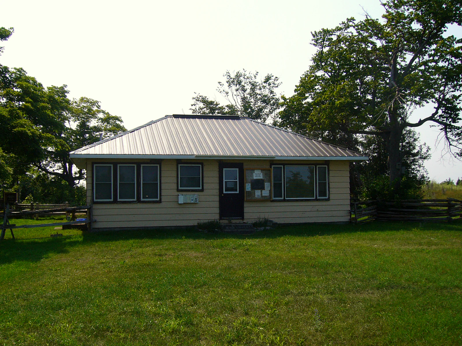 A single-story, light-colored building with a metallic roof and dark window frames situated in a grassy clearing with trees.