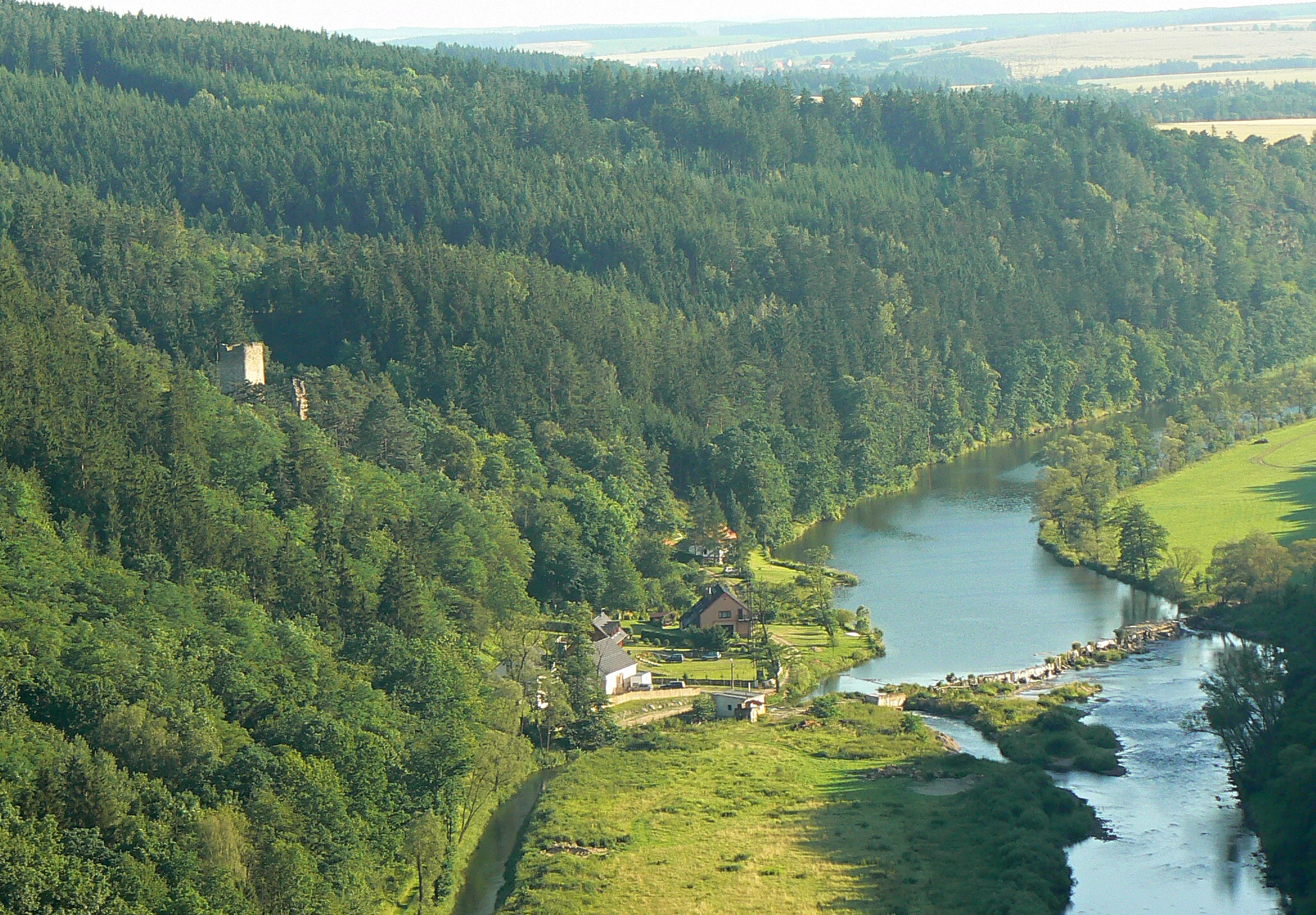 An aerial view of a river bending through a deep, tree-covered valley, with small structures visible on the riverbank and a ruin situated on the forested slope.