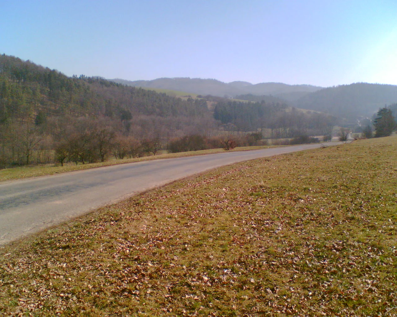 An asphalt road curves through a rural landscape with sparse, dry grassy fields in the foreground and forested hills in the distance under a clear, bright sky.
