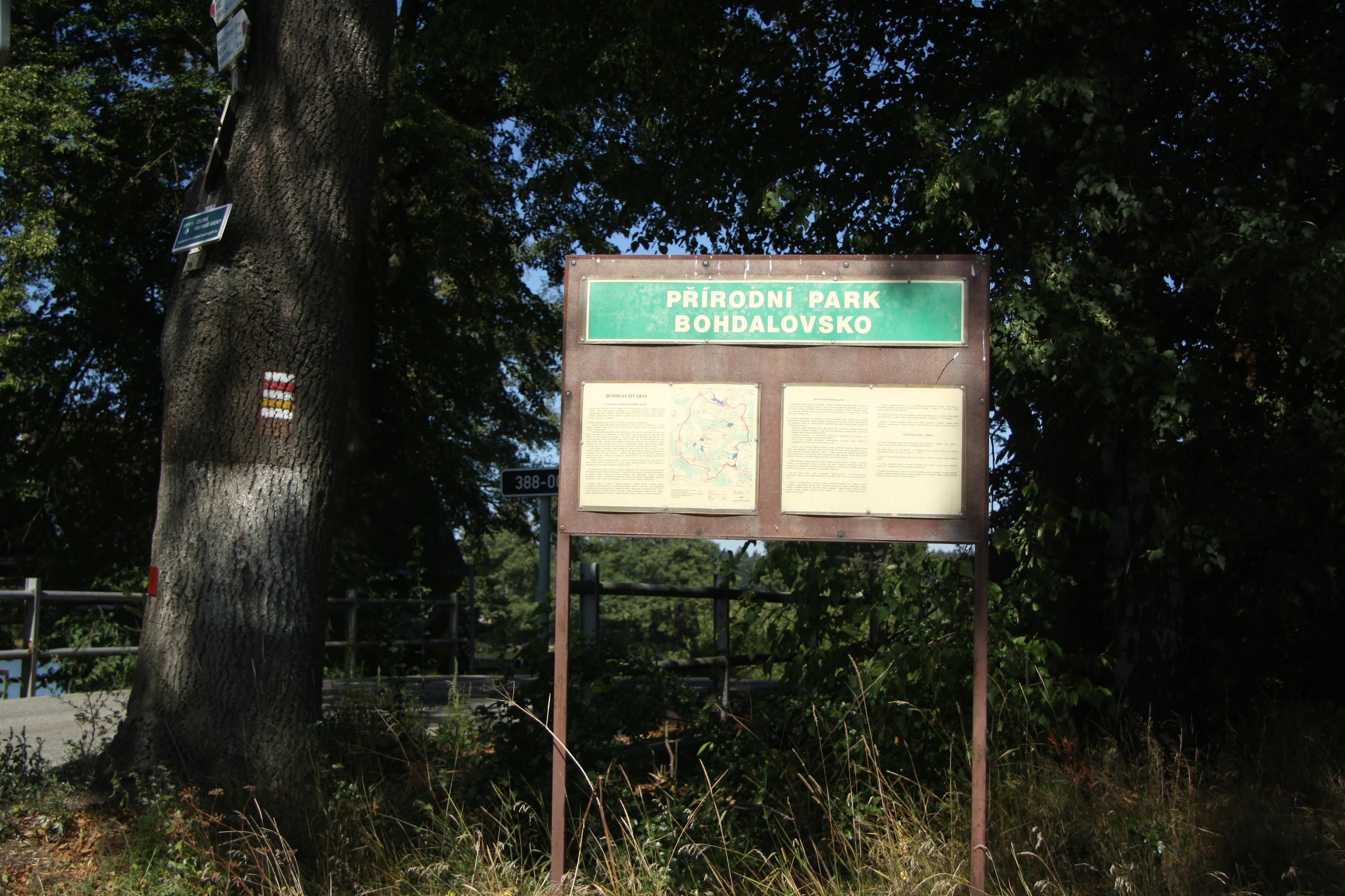 A brown-framed rectangular information board for Přírodní park Bohdalovsko standing in a wooded area with a tree to the left bearing trail markers.
