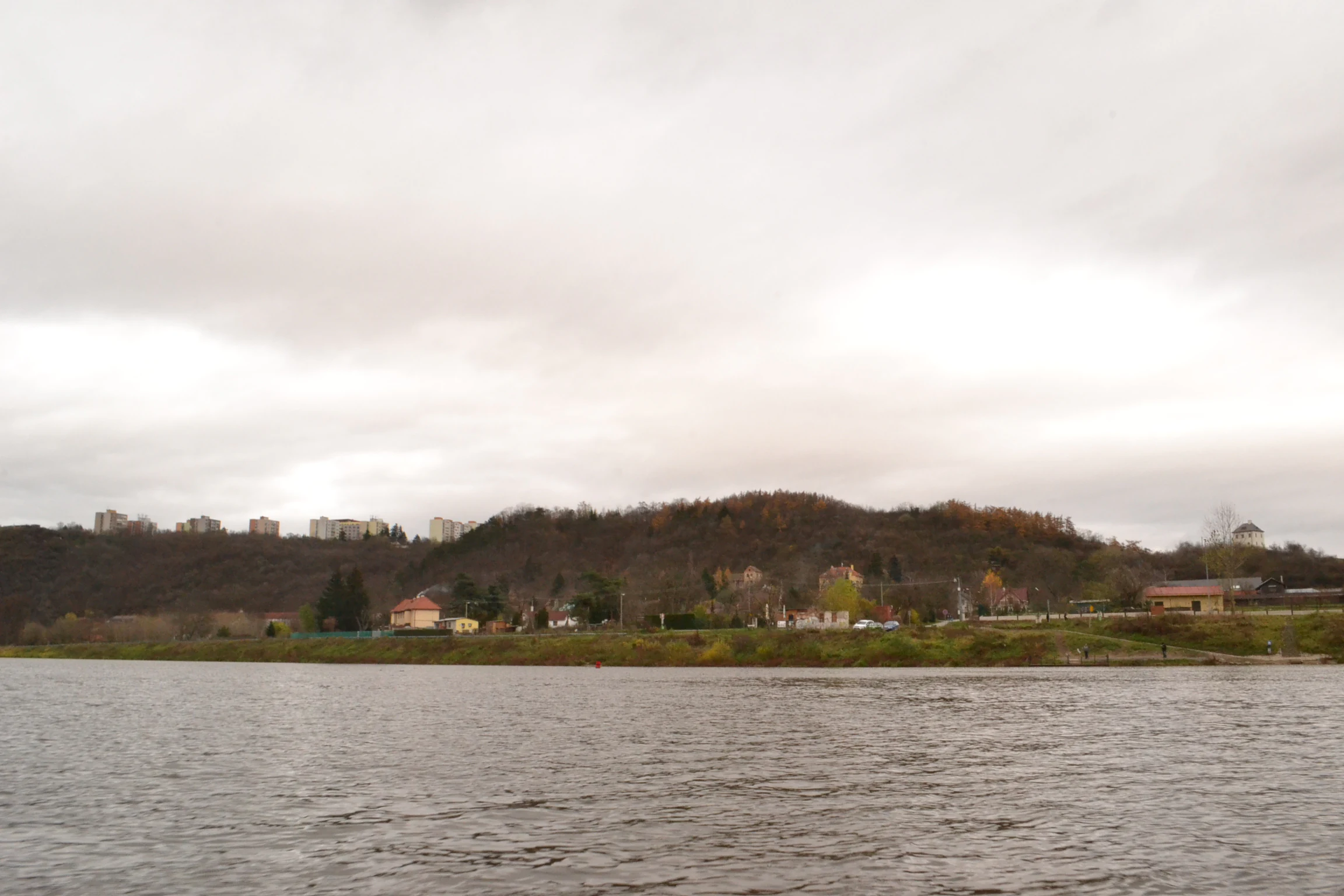 A wide shot looking across the Vltava River toward a forested hill and residential structures in the Podhori district under an overcast sky.