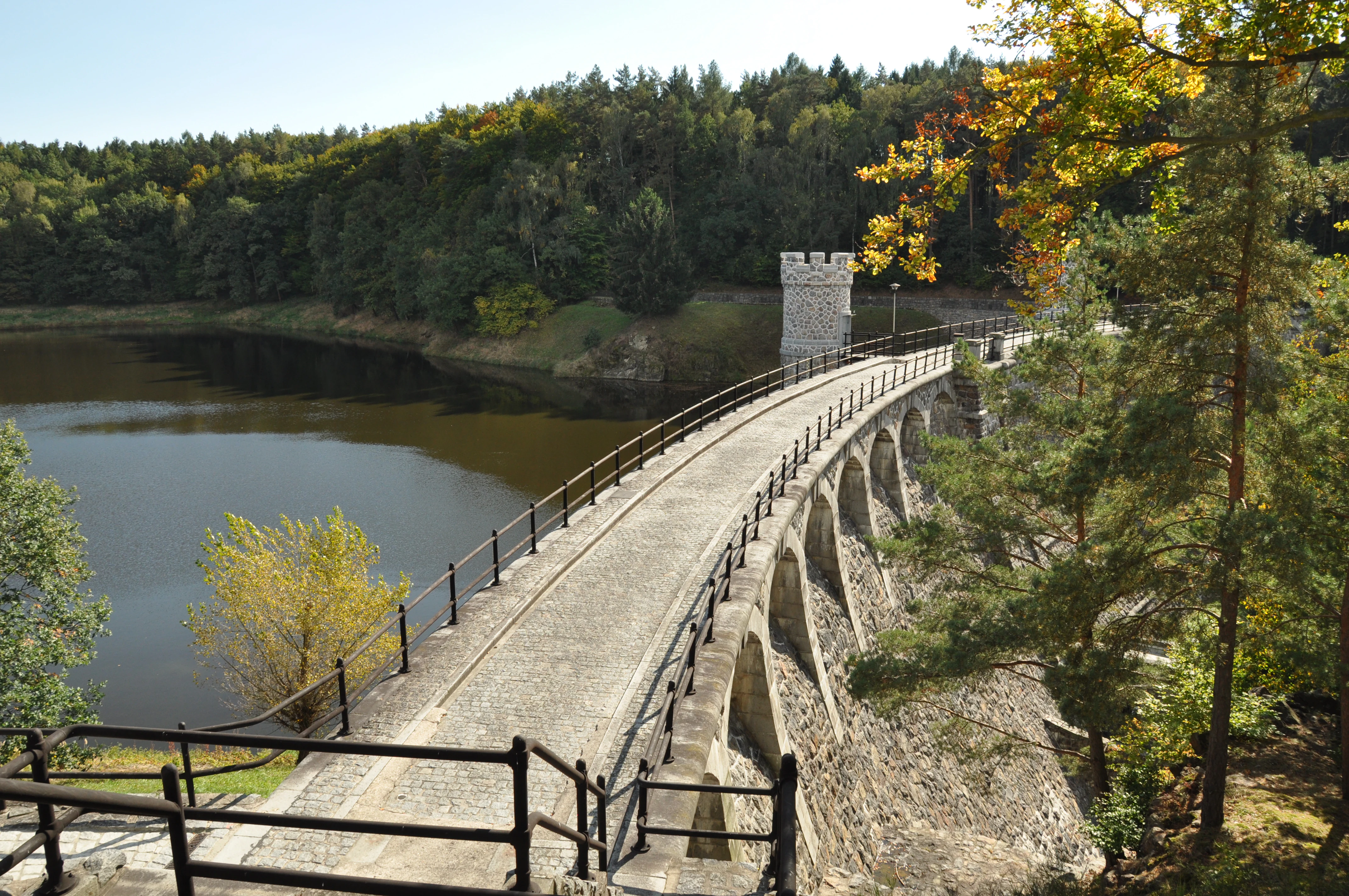 A wide angle view of the Pařížov dam, featuring a curved stone wall with arched supports and a crenelated tower, set within a forested valley with a calm reservoir on the left.
