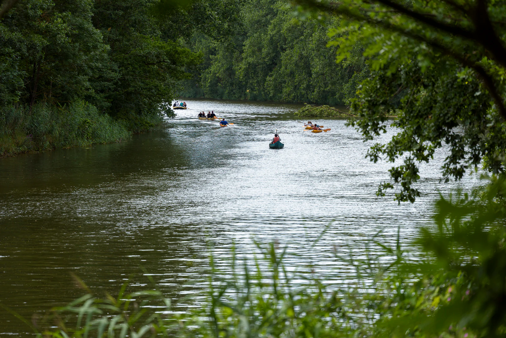 A wide river flows through a dense green forest with several people paddling in small boats and rafts.