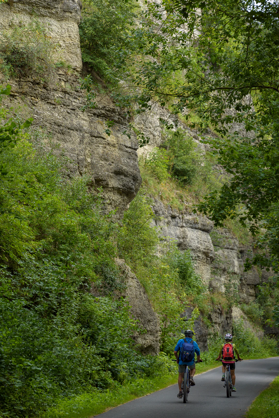 Two cyclists riding away on a paved path next to a steep, vegetation-covered rocky cliff.