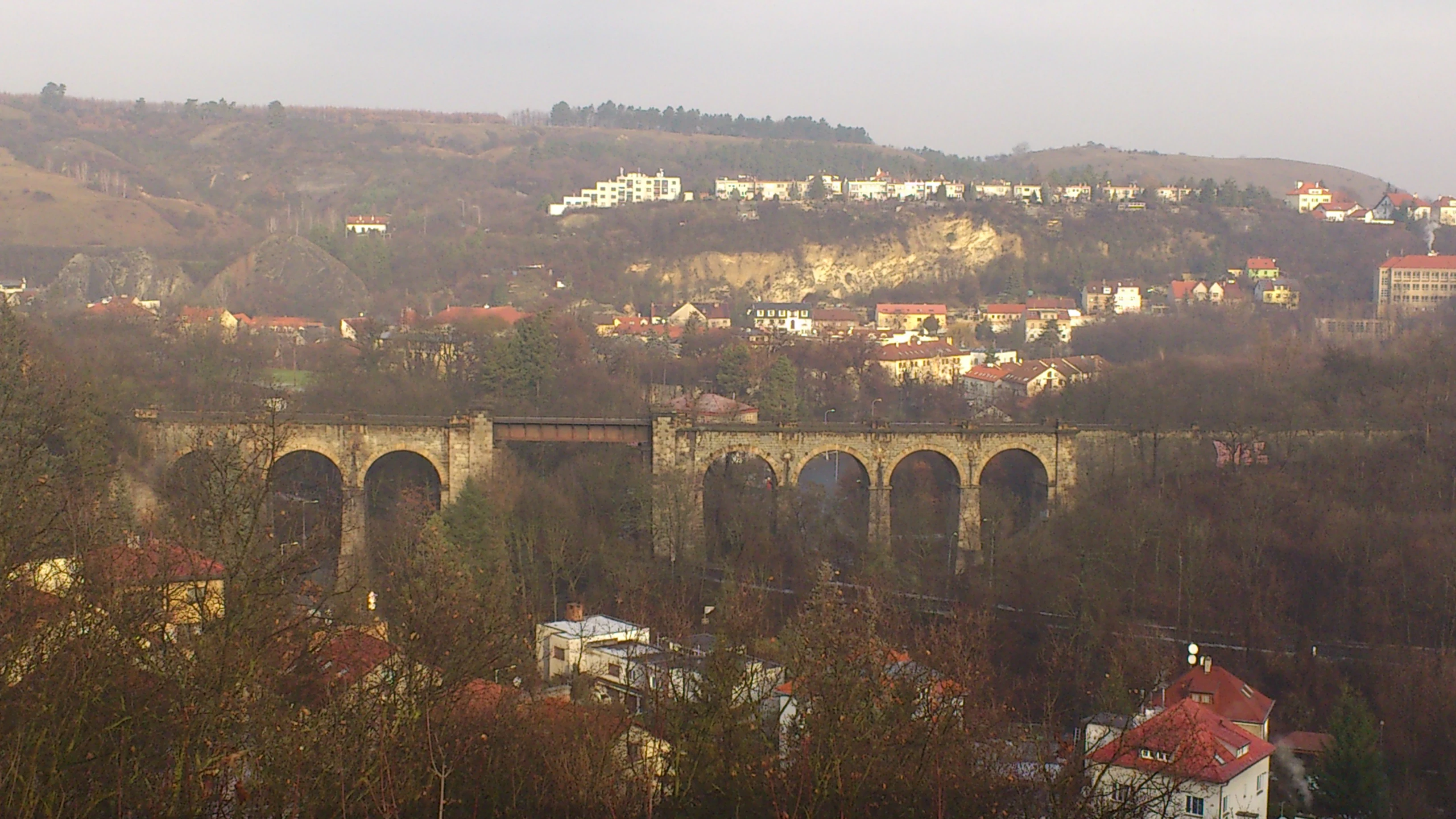 A wide shot of a stone arch railway viaduct spanning a wooded valley in the Prokop Valley, with suburban housing visible on the hillside above.