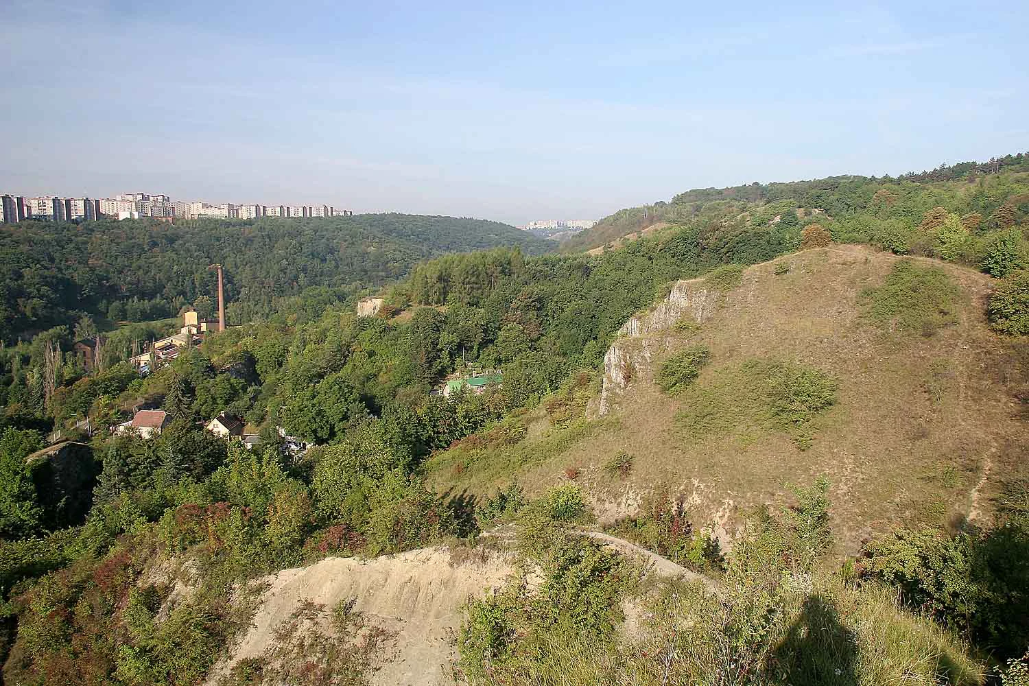 Panoramic view of the rolling green hills and rocky terrain of Prokopské údolí valley in Prague, with a residential housing estate visible on the distant ridge.