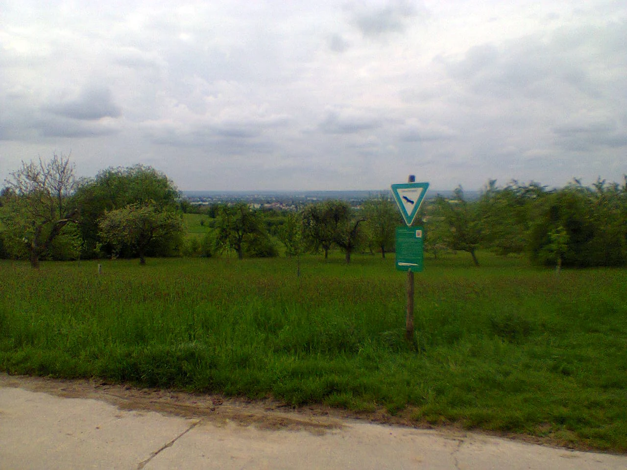A nature reserve sign with a green triangle symbol stands in a grassy field with trees under a cloudy sky.