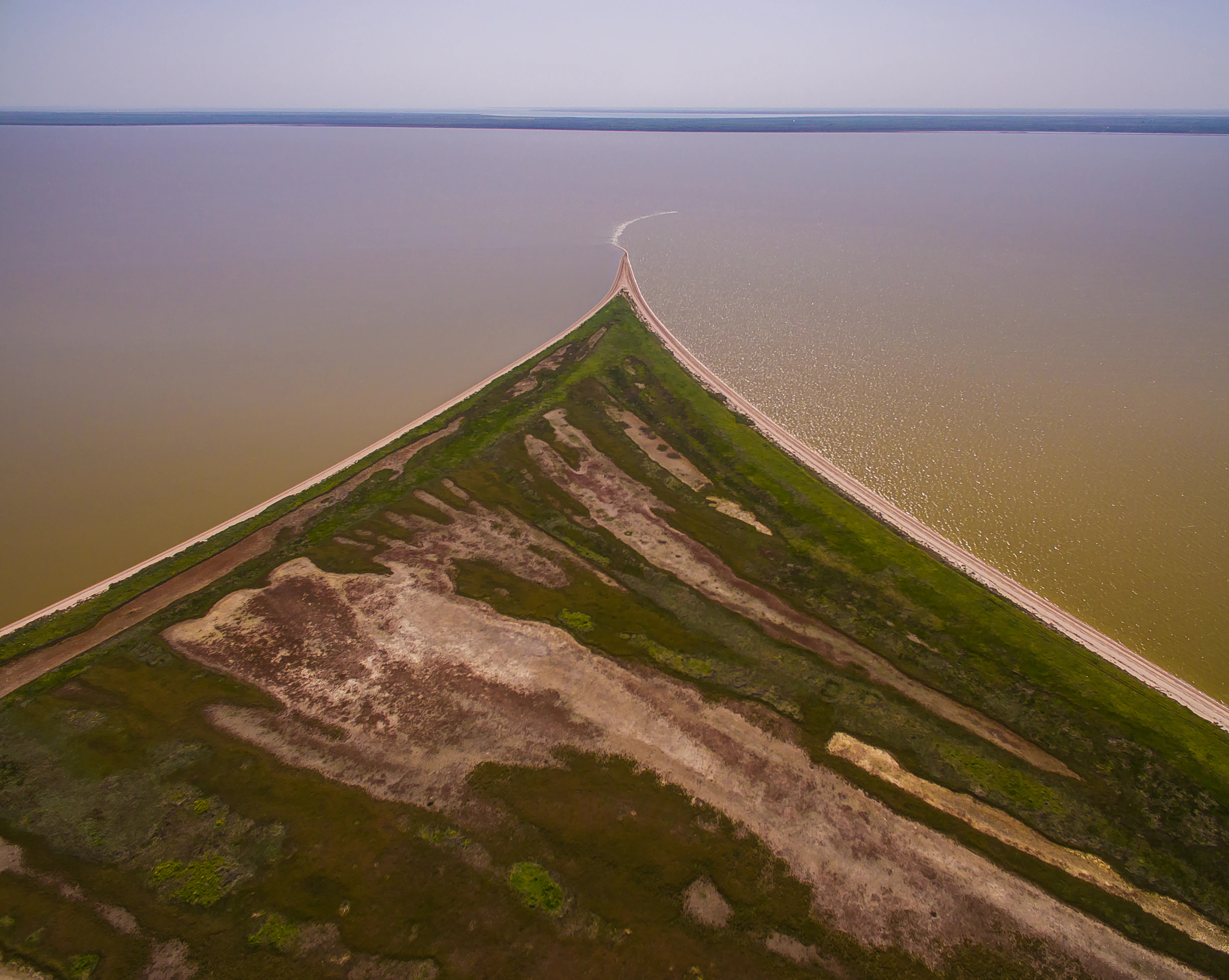 Aerial view of a narrow coastal spit extending into the sea, with sandy and grassy terrain, and calm waters on both sides