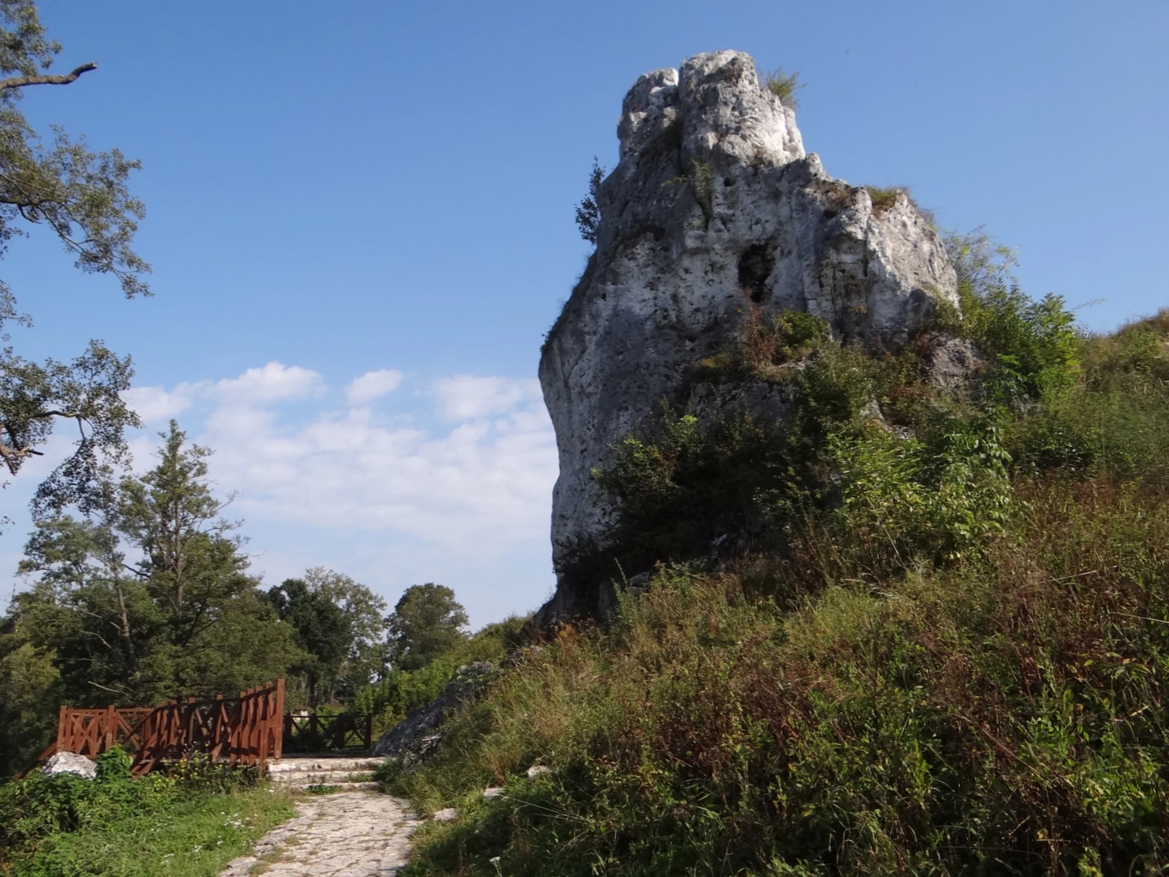 Una gran formación rocosa de piedra caliza y dentada que se eleva sobre una colina cubierta de hierba, junto a un sendero de piedra y una pequeña valla de madera.