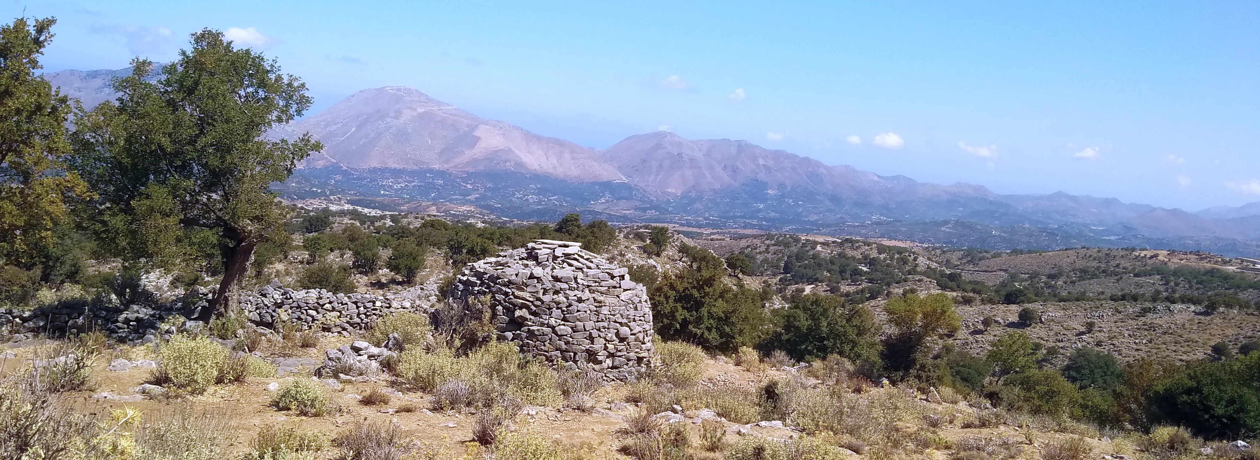 Una estructura circular de piedra, posiblemente una choza de pastor, se encuentra en un paisaje montañoso escarpado y rocoso bajo un cielo despejado, con cadenas montañosas distantes visibles.