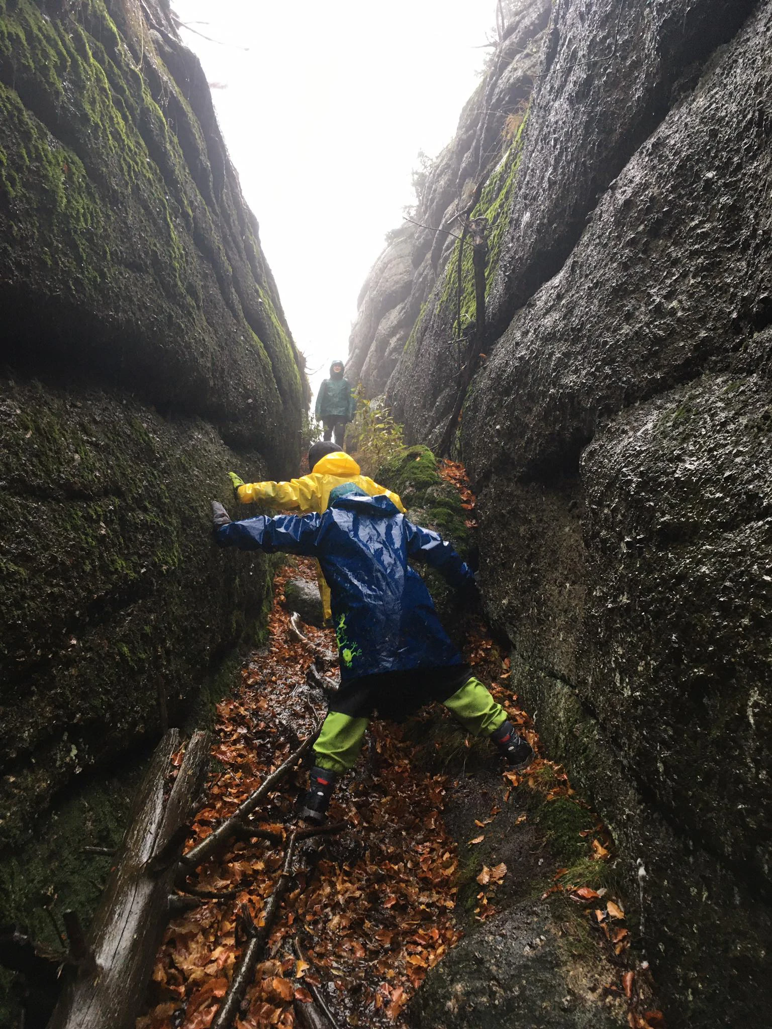 Two people in colorful rain jackets carefully climbing through a narrow, moss-covered granite chasm covered in fallen leaves.