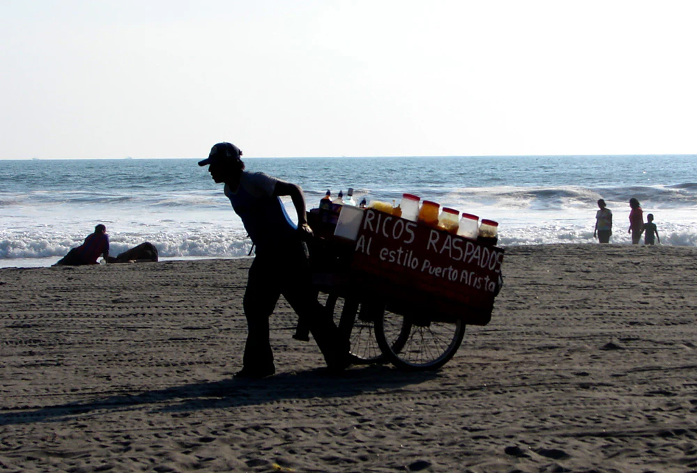 A person pushing a cart with a sign that says RICOS RASPADOS on a sandy beach, with the ocean and breaking waves in the background.