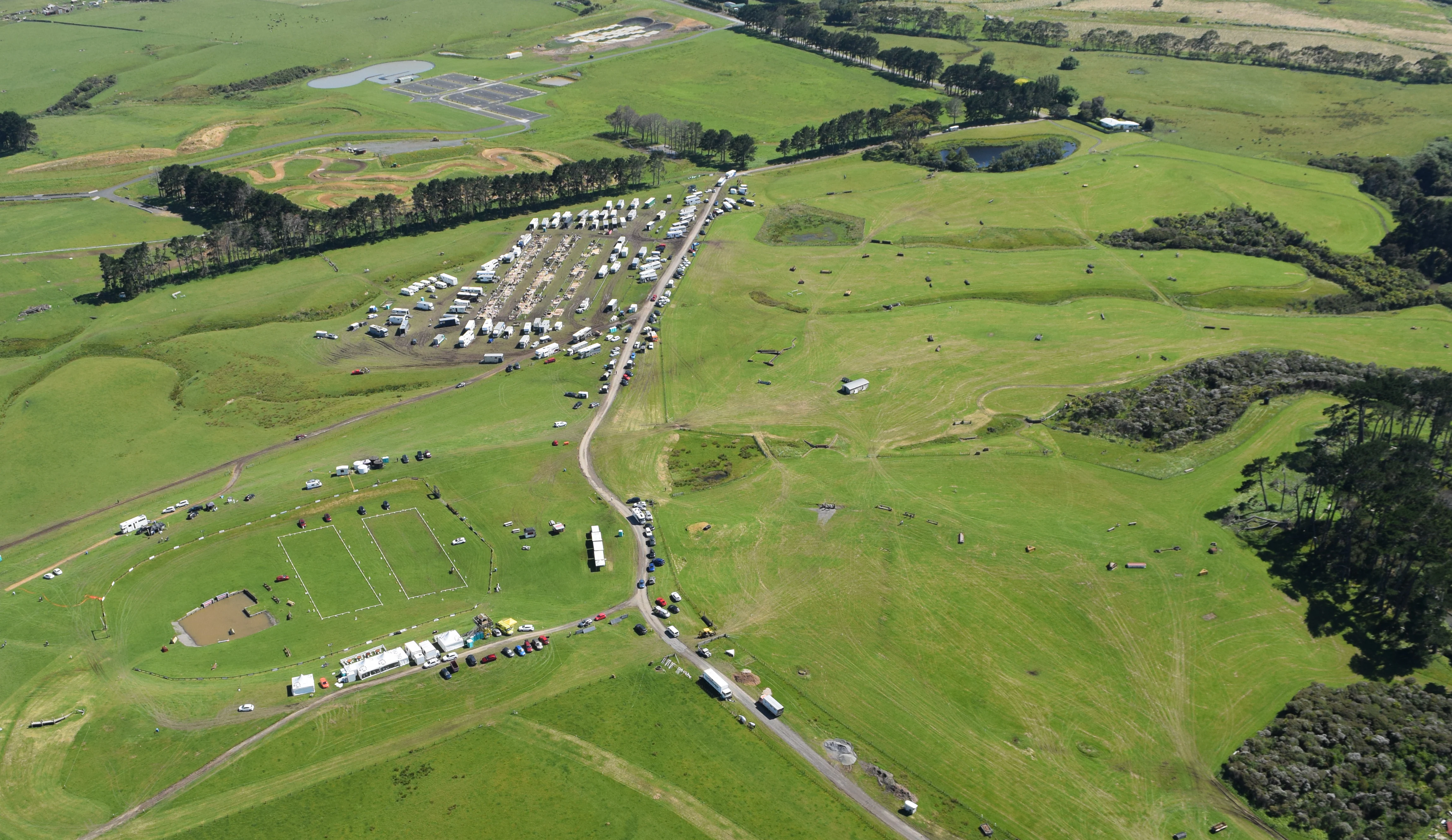 An aerial view showing an expanse of rolling green pasture in Puhinui Reserve, with dirt roads, parking areas, and clusters of vehicles and temporary structures.