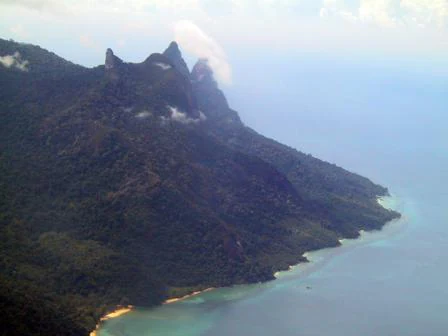 An aerial view of the mountainous, heavily forested coast of Tioman Island, showing jagged peaks and the meeting of dense forest and turquoise sea.