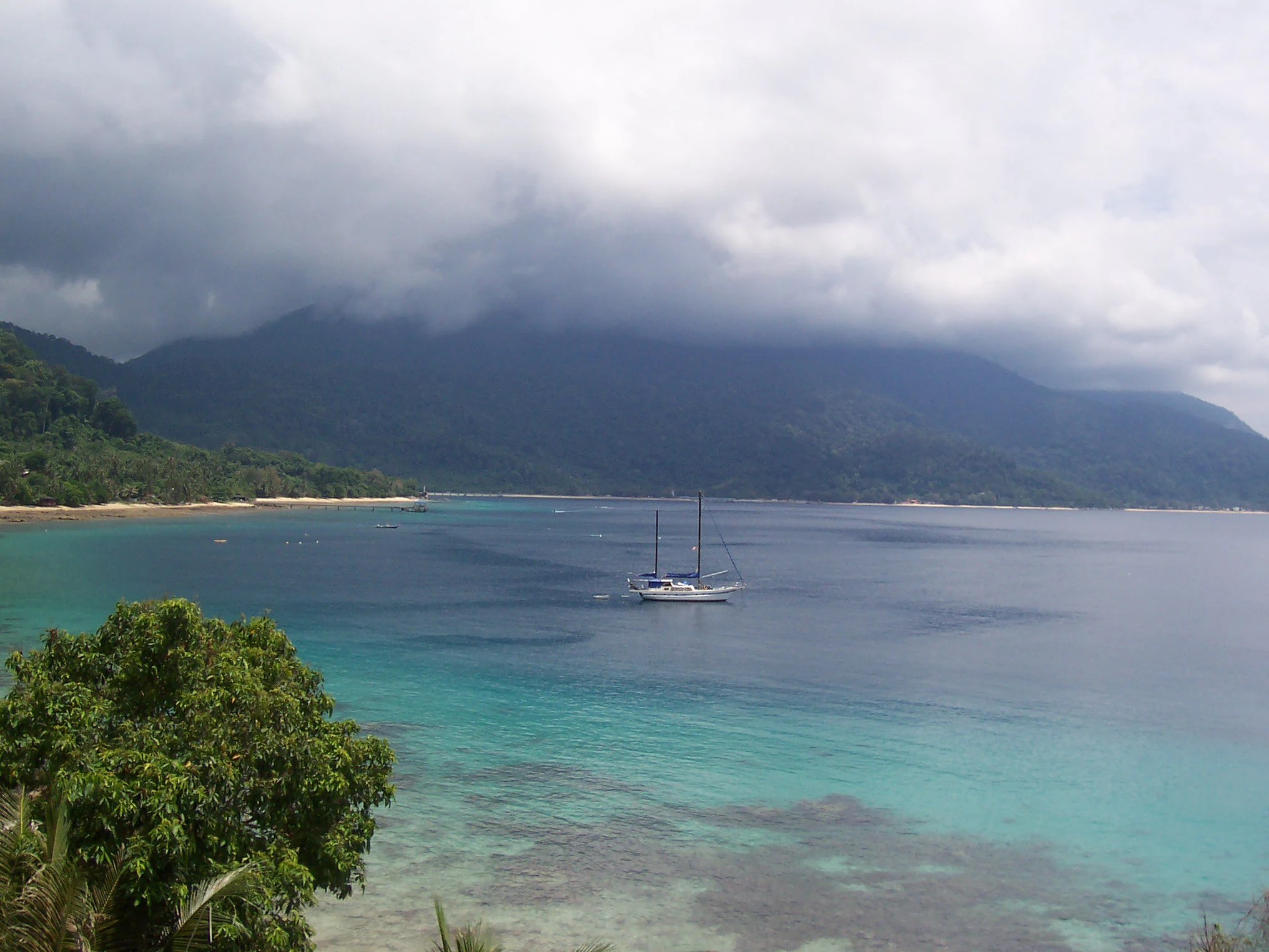 A wide view of a calm tropical bay with turquoise water in the foreground; a sailboat is anchored in the center; a forested coastline and hazy mountains are in the background under a cloudy sky.