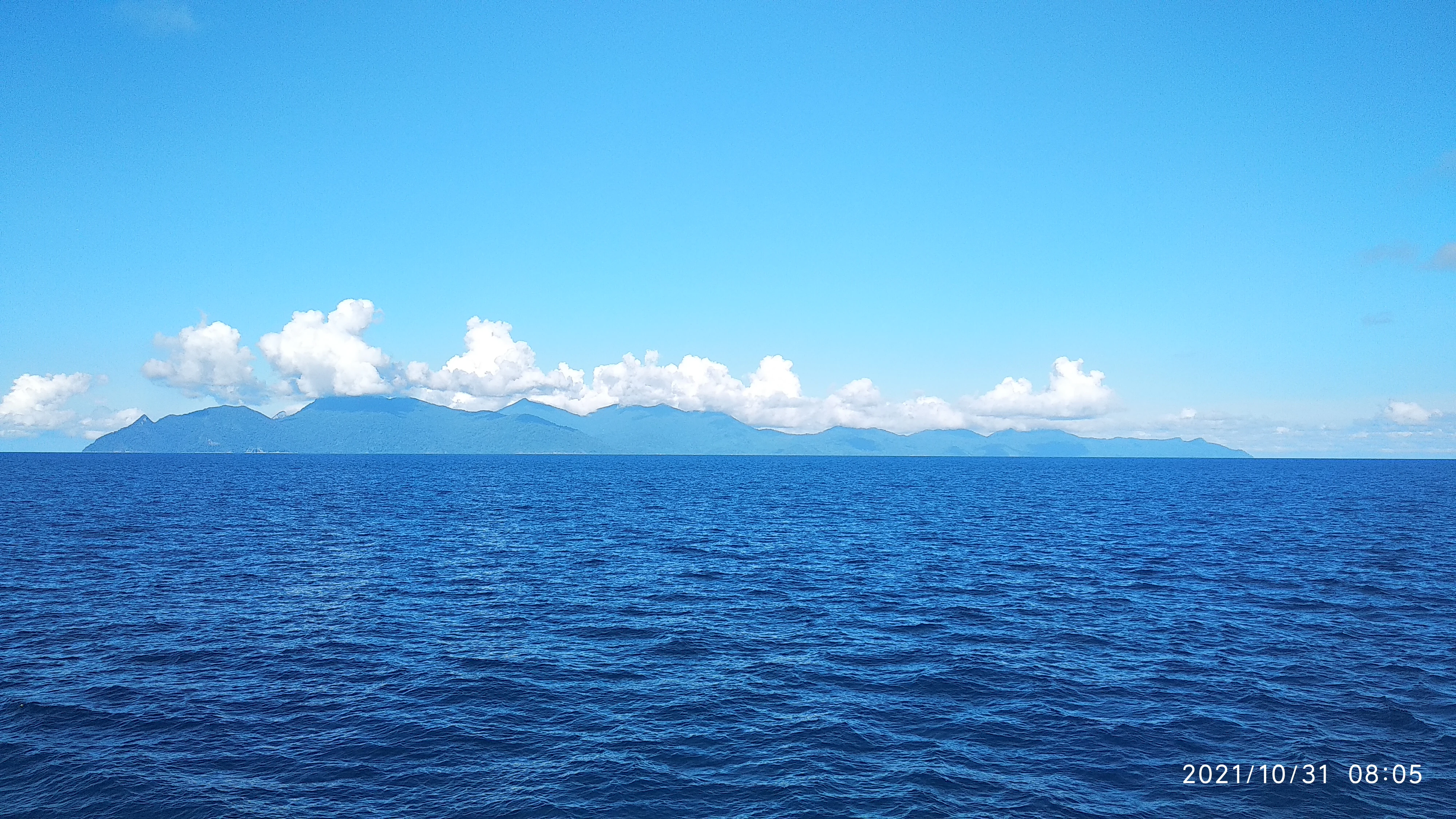 A wide, eye-level shot of a large, mountainous island silhouette against a clear blue sky, viewed from across the open ocean.