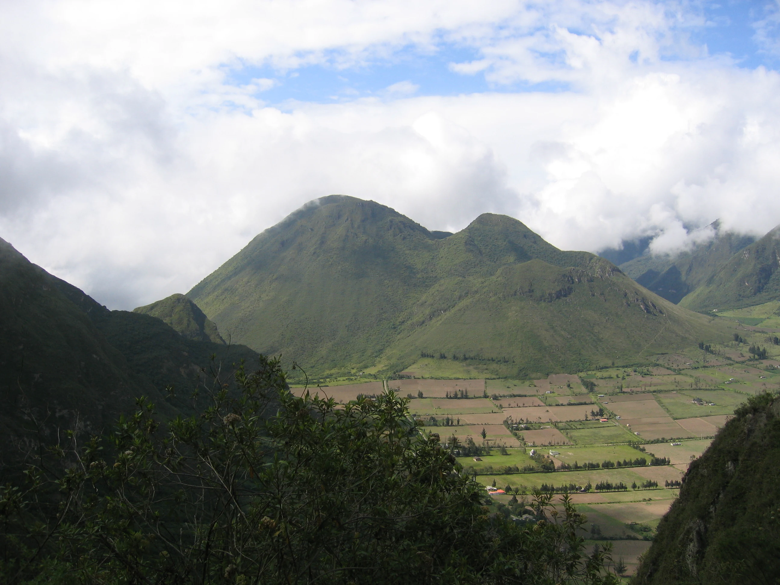 A wide angle landscape view of a green volcanic caldera with agricultural fields on the crater floor, looking towards the Pondoña and Rumiloma volcanic domes under a cloudy sky.