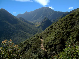 A dirt trail winds down a steep, green mountain slope into a large volcanic caldera with mountain peaks in the distance.