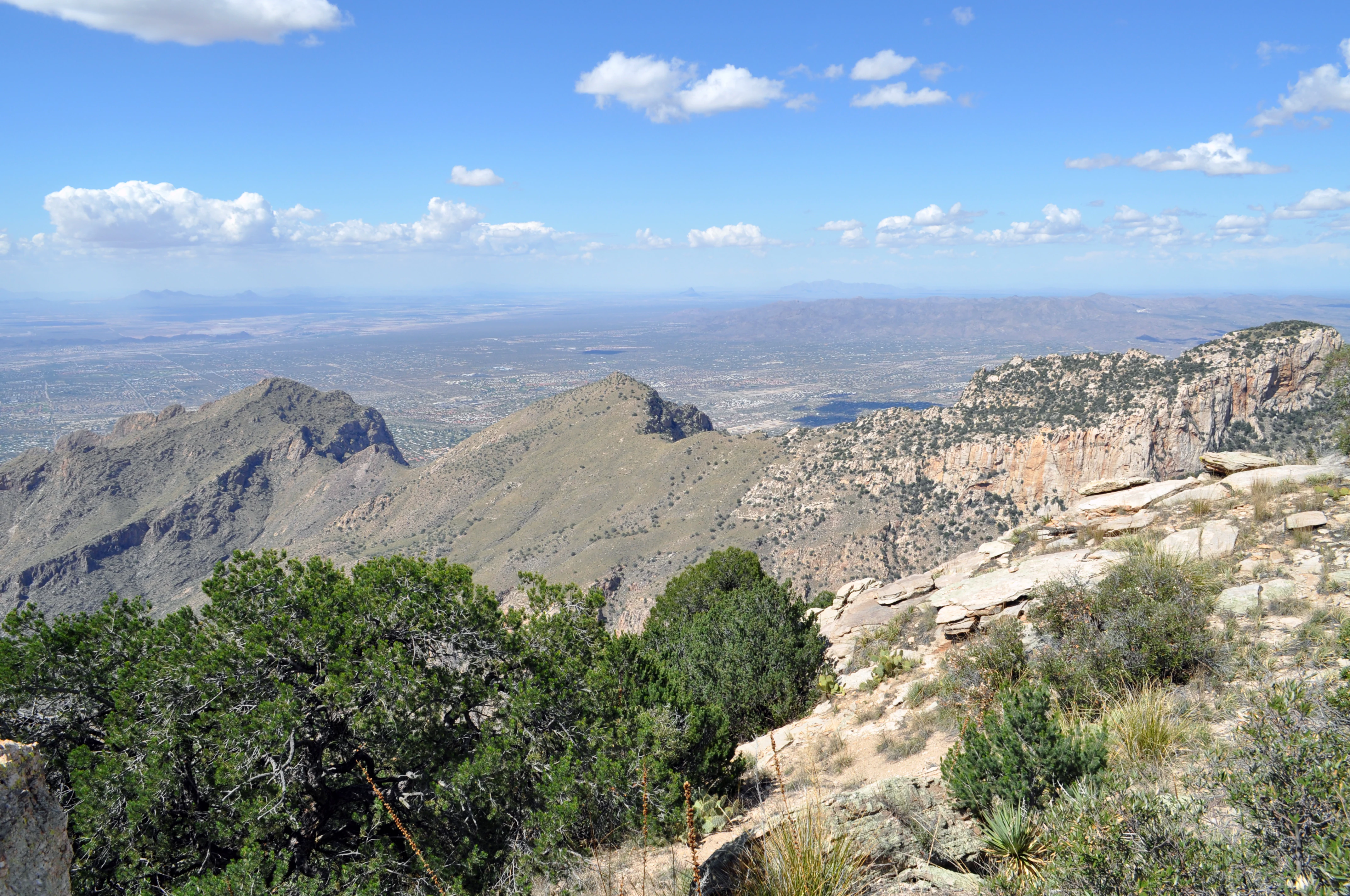 A high-angle scenic view of the rugged Pusch Ridge mountains under a clear blue sky, with rocky foreground slopes and a distant desert valley.