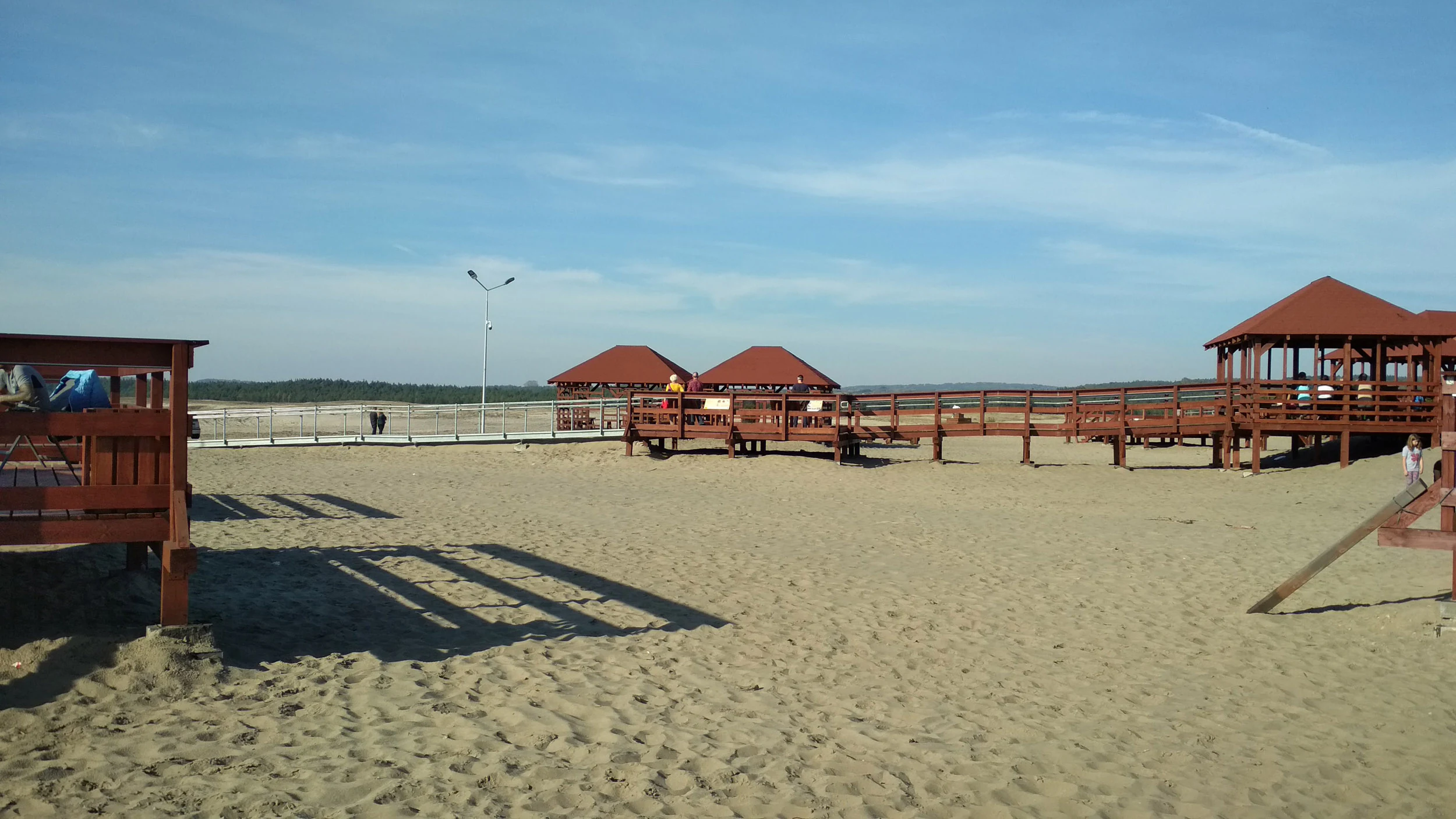 A sandy landscape features wooden raised observation platforms and walkways with red roofs at Pustynia Błędowska.