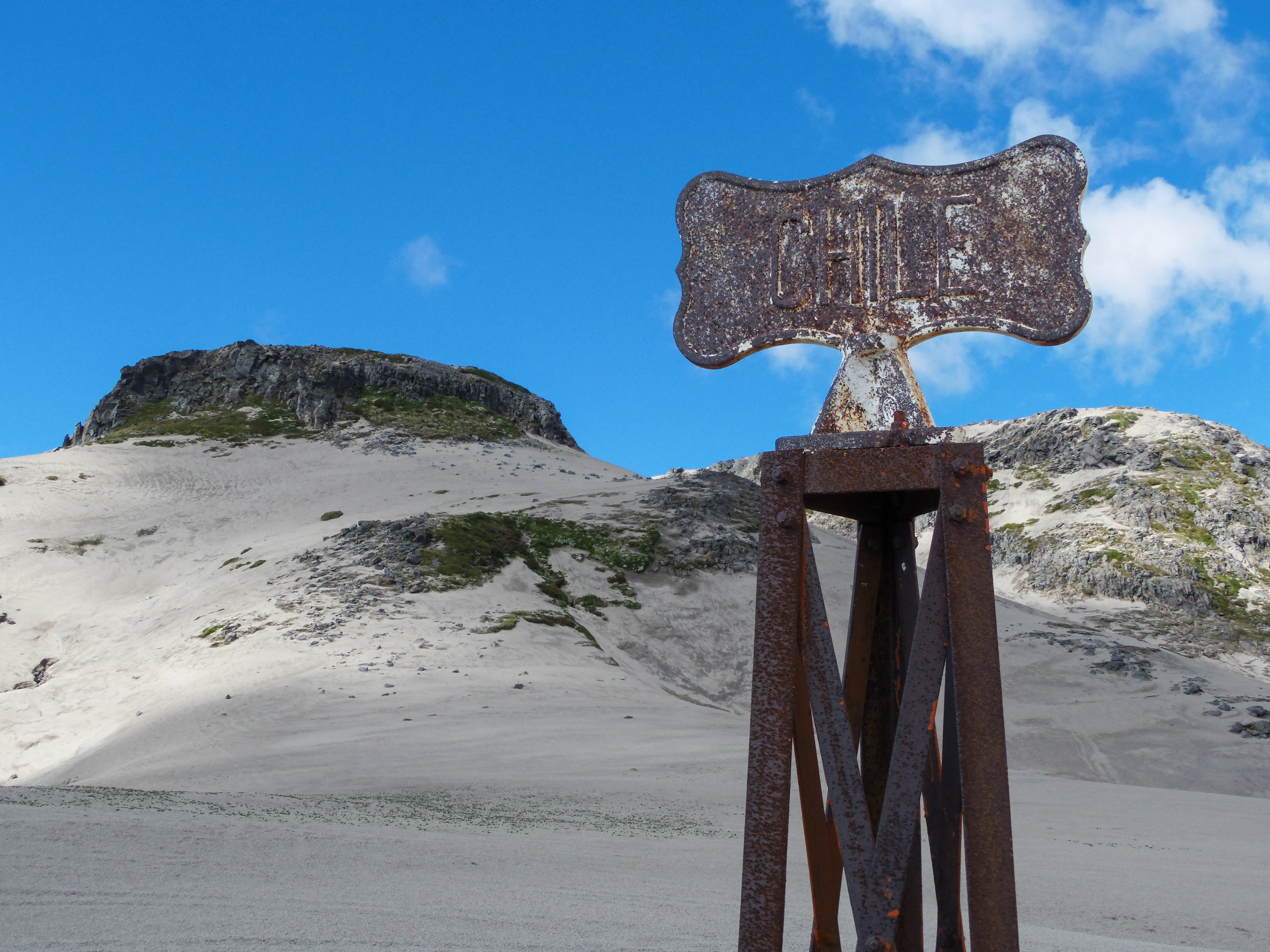 Rusty metal sign structure with faded text on a stand, set against sandy terrain with rocky hills in the background under a blue sky with clouds