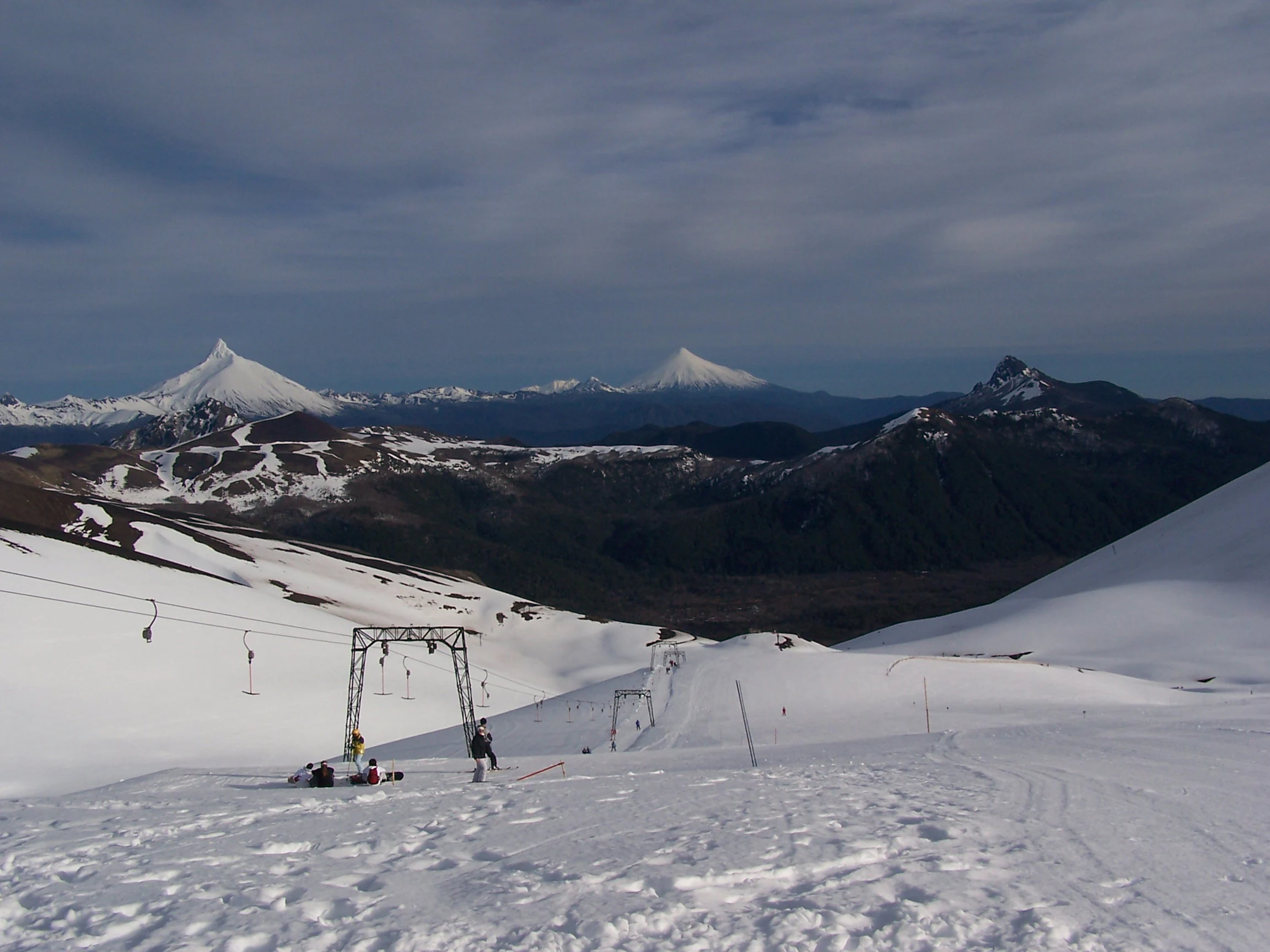 Snow-covered ski slope with ski lift structure, two people on the slope, and mountain peaks in the distance under a cloudy sky