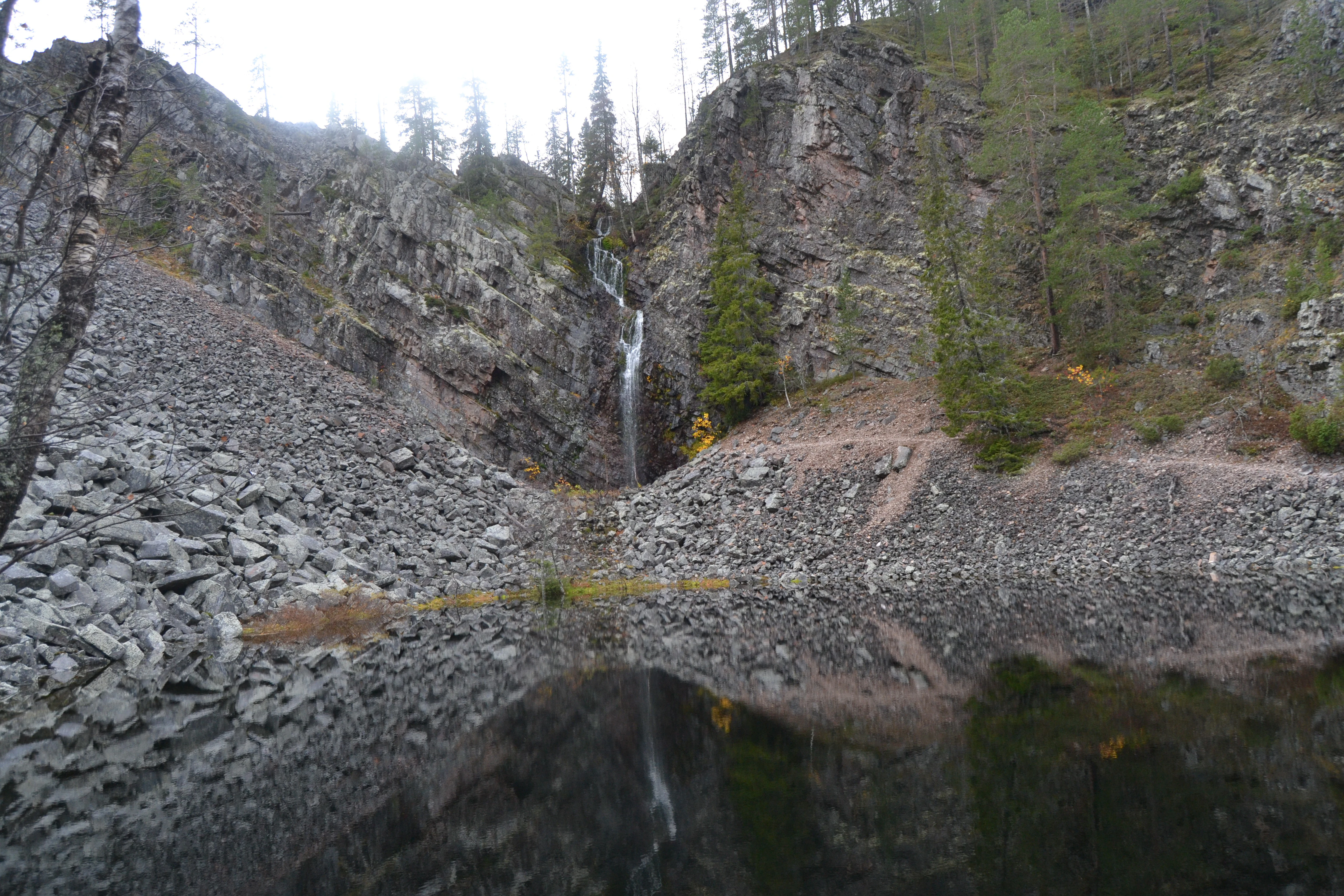 Waterfall cascading down a rocky cliff face with a small body of water reflecting the waterfall and surrounding landscape