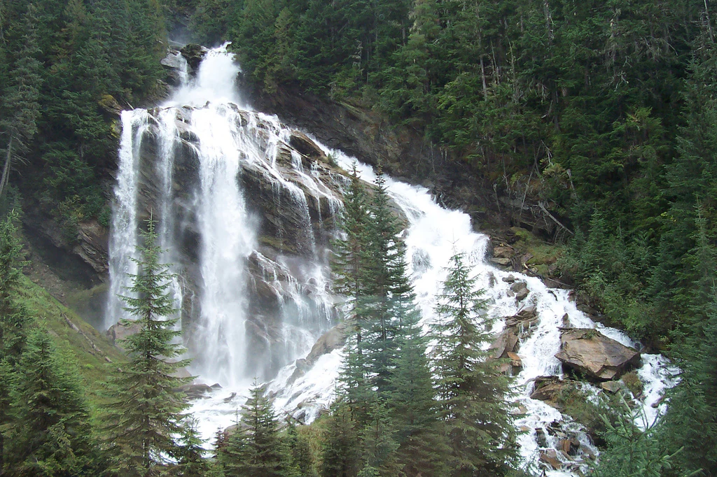 A wide, tiered waterfall flows down a rocky cliff face surrounded by dense evergreen forest.