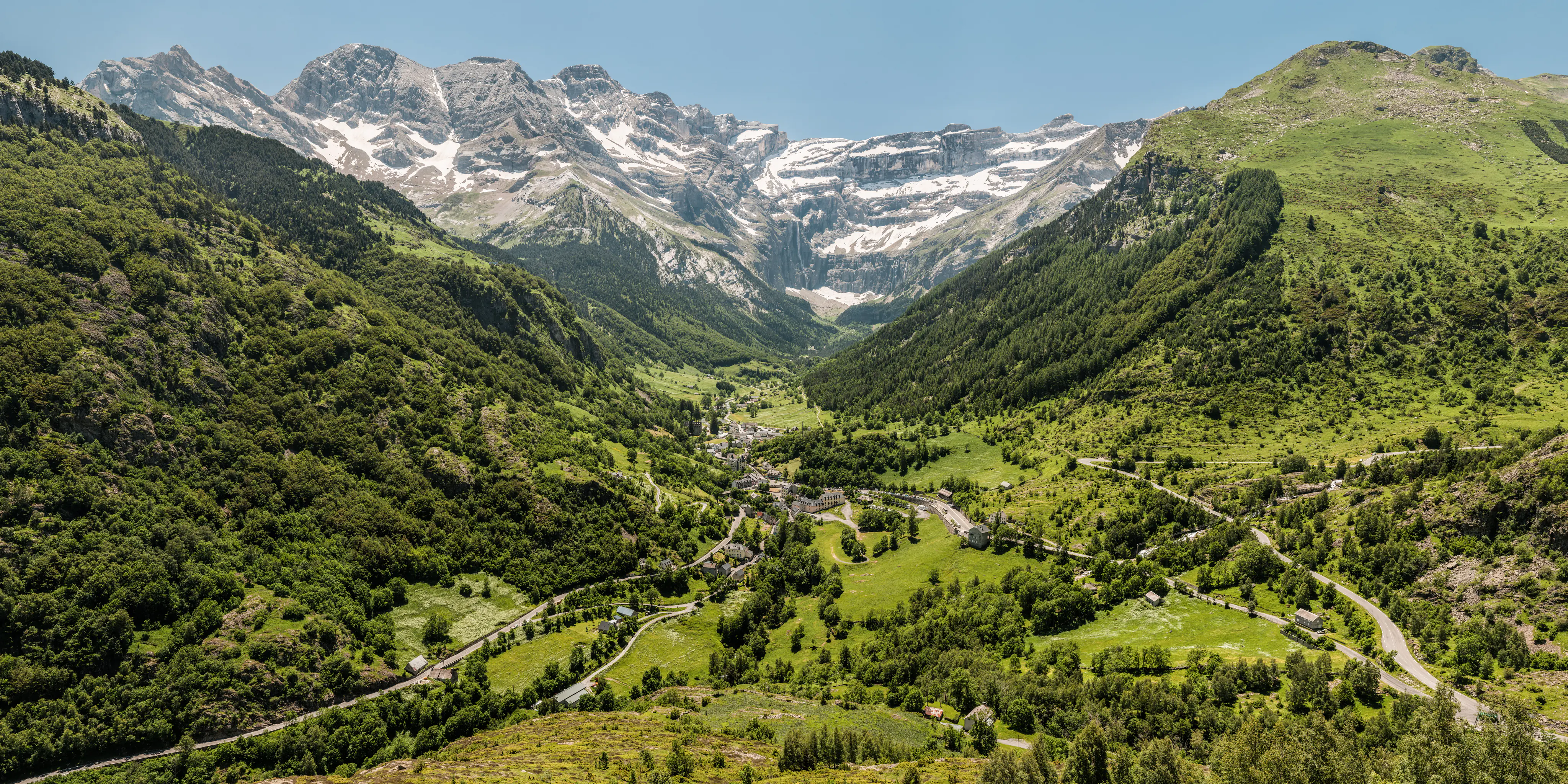 A wide landscape view of the Cirque de Gavarnie showing a deep green valley with a small village nestled in the center; the valley is surrounded by steep, snow-dusted, jagged limestone mountain peaks under a clear blue sky.