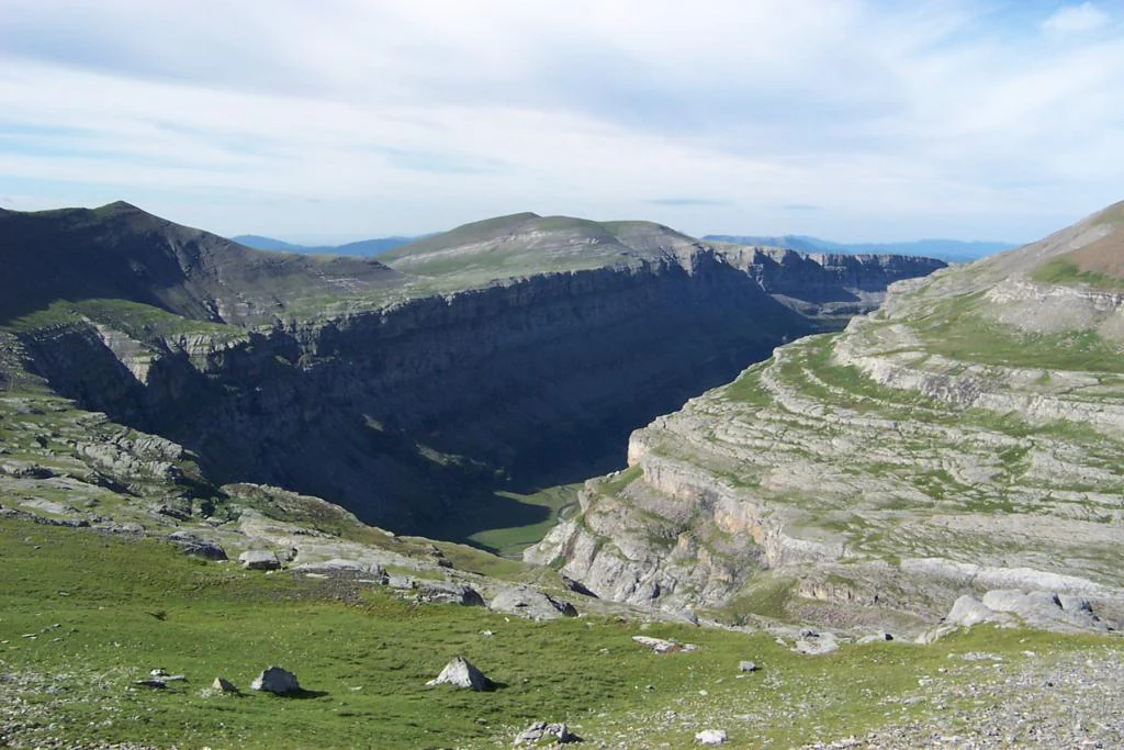 A high-angle view looking down into the deep, rocky Ordesa Valley canyon with layered limestone cliffs on both sides and grassy, sloping terrain in the foreground.