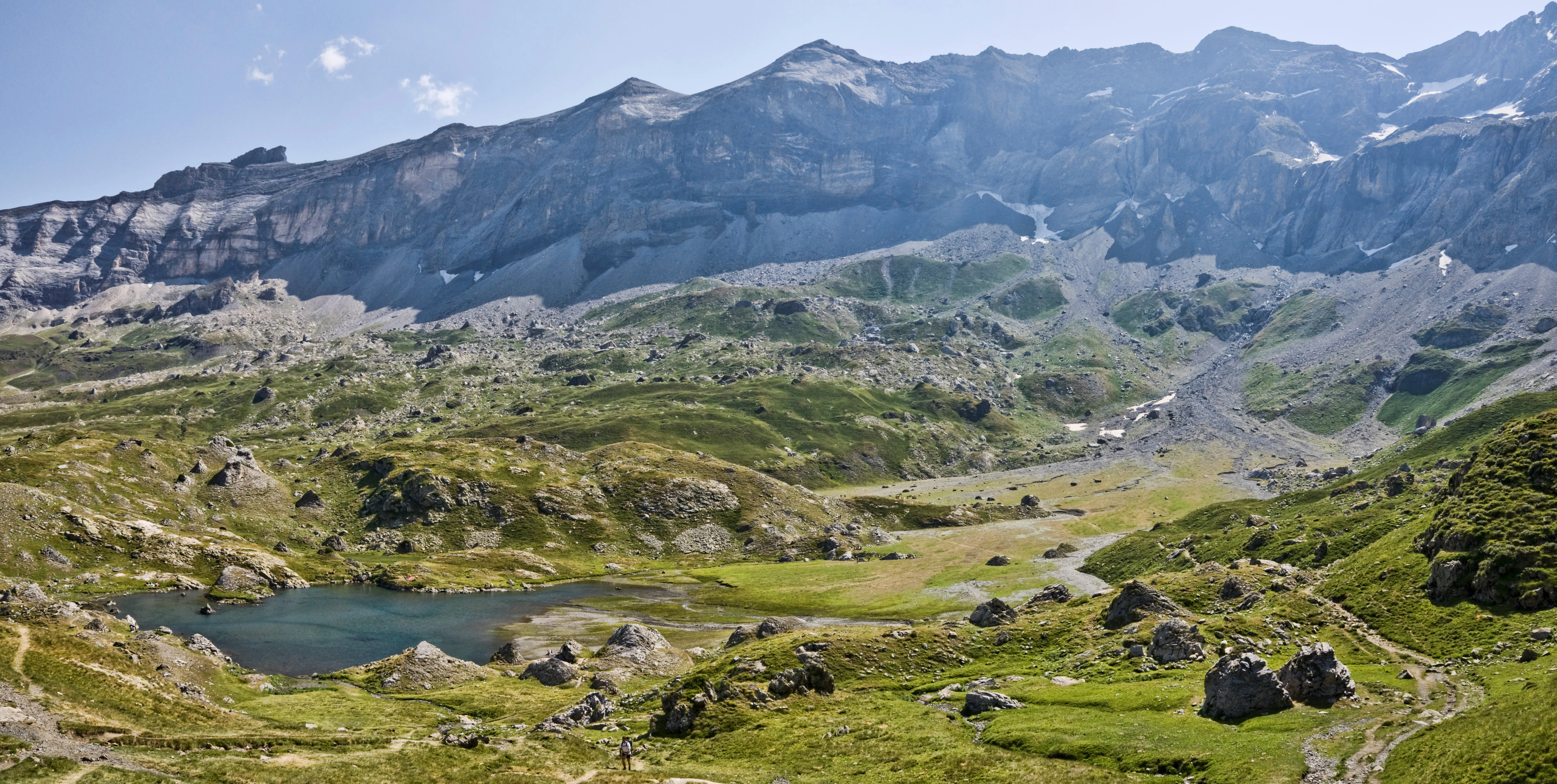 A panoramic landscape view of Cirque de Troumouse in the Pyrenees, featuring a large, grassy glacial cirque bowl with a small lake in the foreground and steep rocky cliffs in the background under a blue sky.