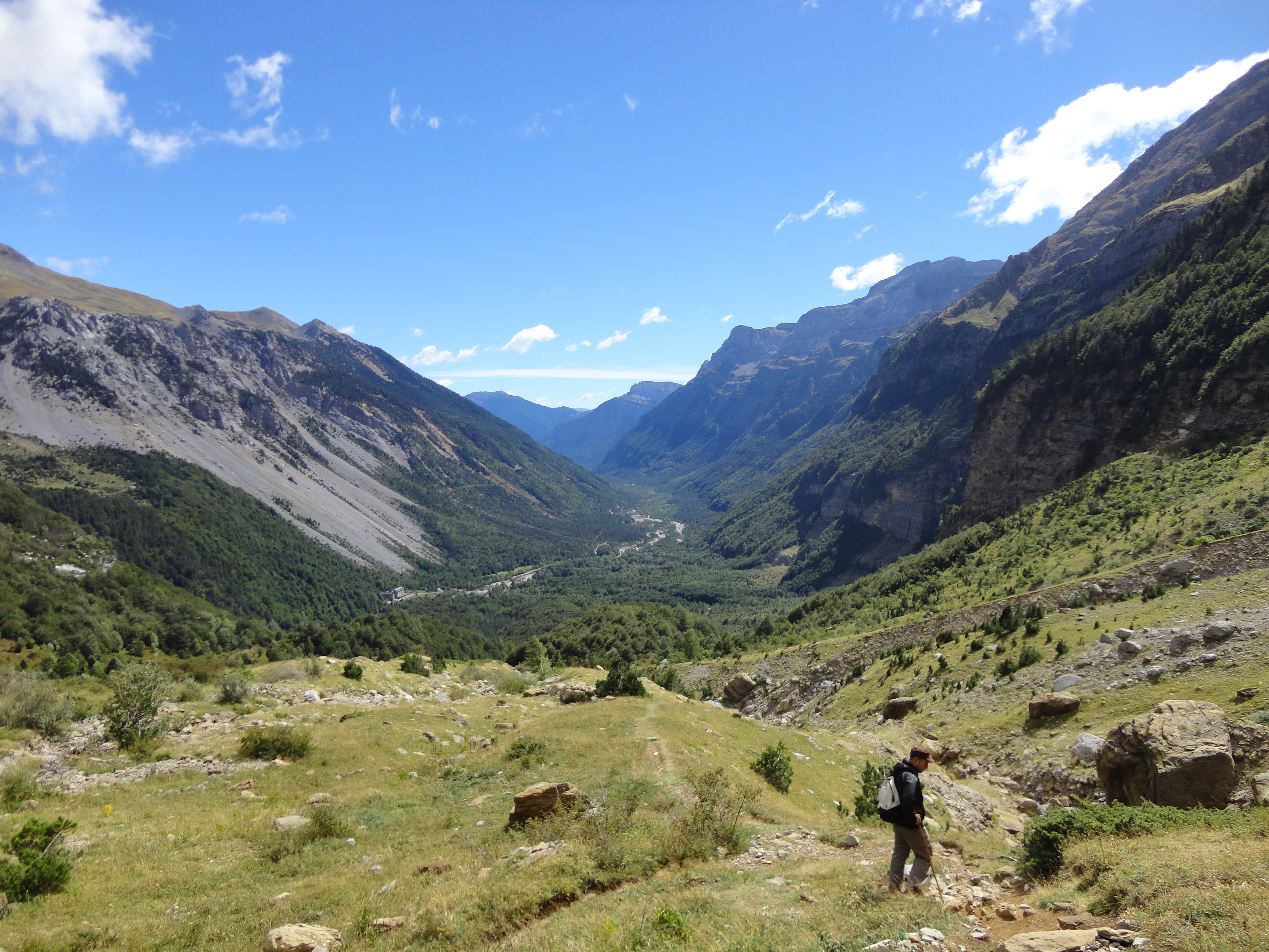 A wide view of a deep, green alpine valley framed by towering, steep mountains under a blue sky, with a lone hiker in the foreground.