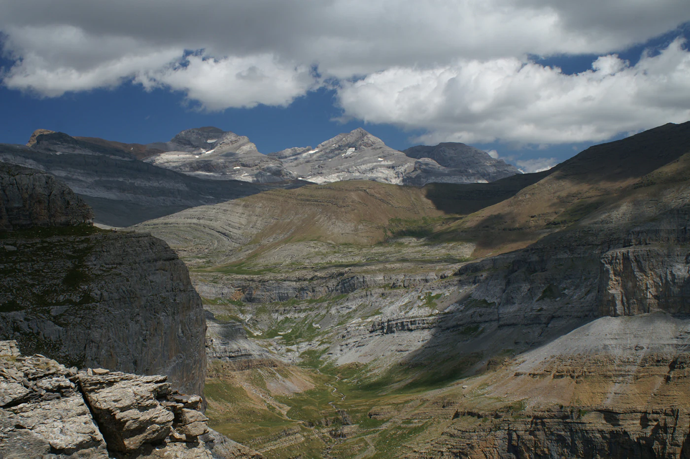 A wide, high-angle view of the rugged, rocky peaks of the Monte Perdido massif under a partly cloudy sky, overlooking the deep, terraced limestone slopes of the Ordesa Valley.