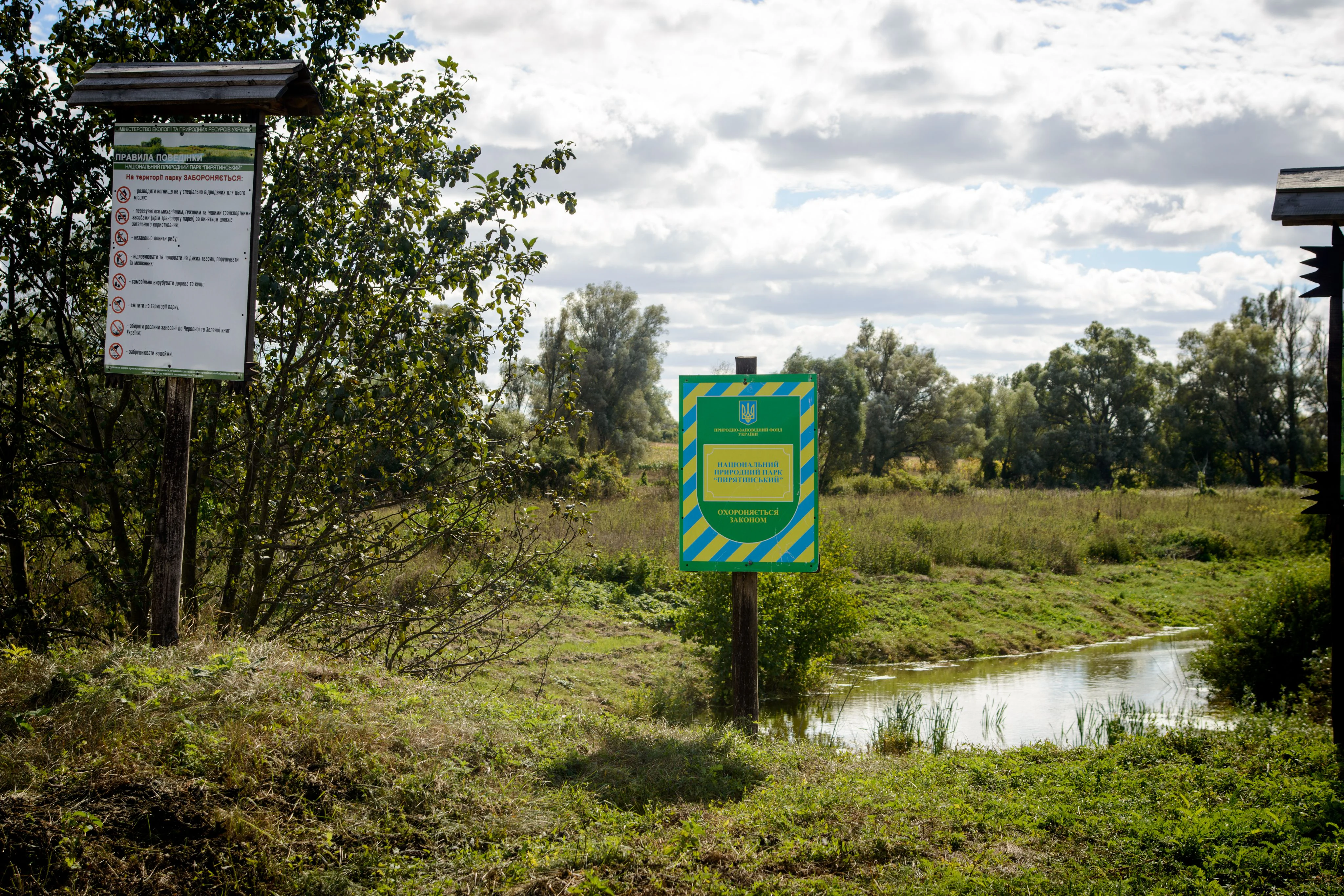 Green and yellow informational sign with text on a wooden post in a grassy area near a river with trees in the background