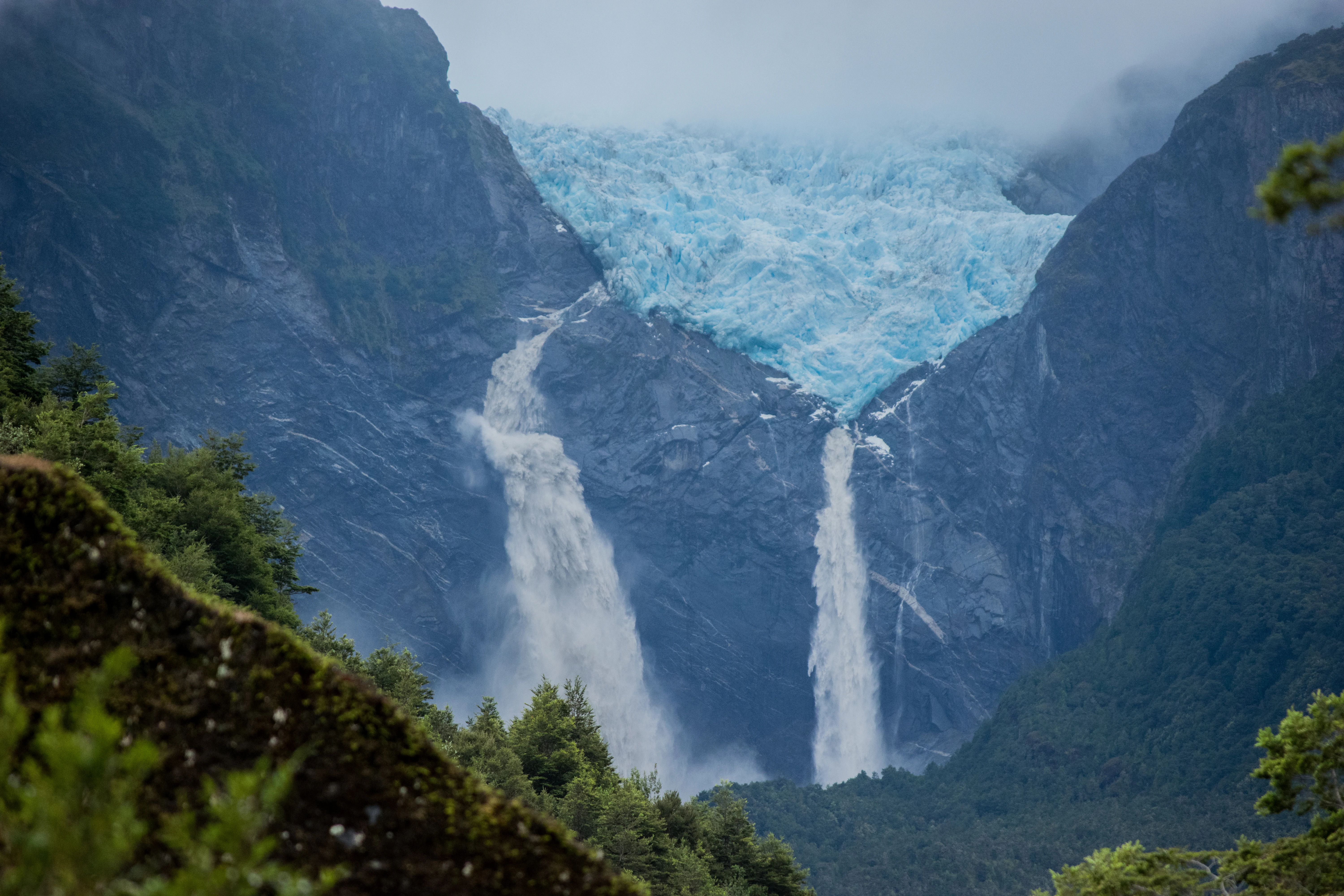A hanging glacier with two waterfalls flowing down steep rocky cliffs, surrounded by dense green forest