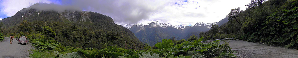 Panoramic view of a winding road through dense green vegetation with snow-capped mountains in the distance, a car on the road, and a person walking