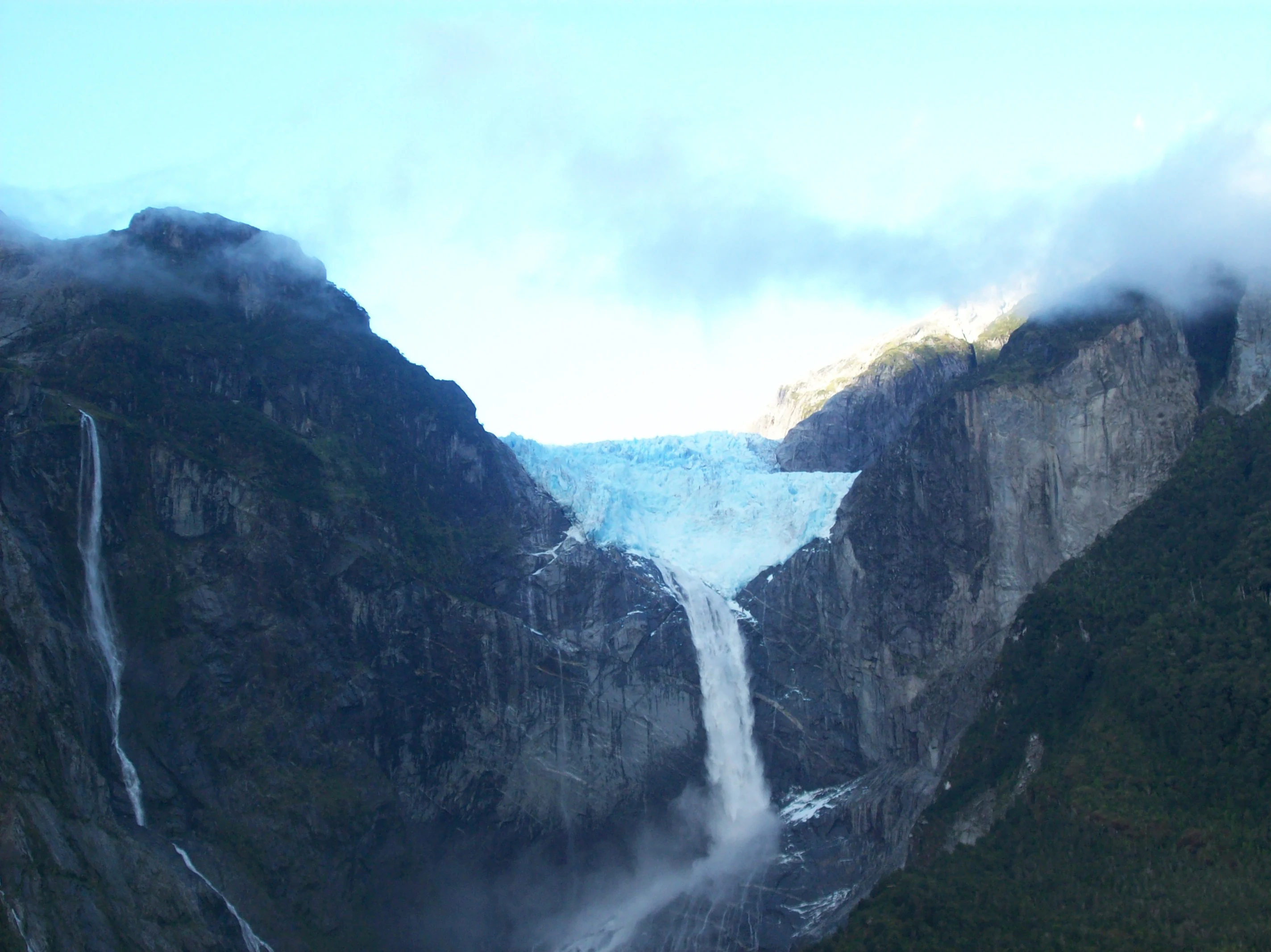 Hanging glacier in Queulat National Park, surrounded by steep rocky mountains with waterfalls cascading down under a partly cloudy sky