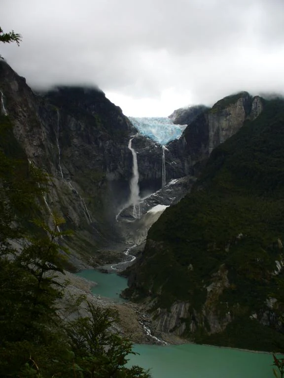Mountainous landscape with a hanging glacier, waterfalls, and turquoise lake surrounded by dense green forests.