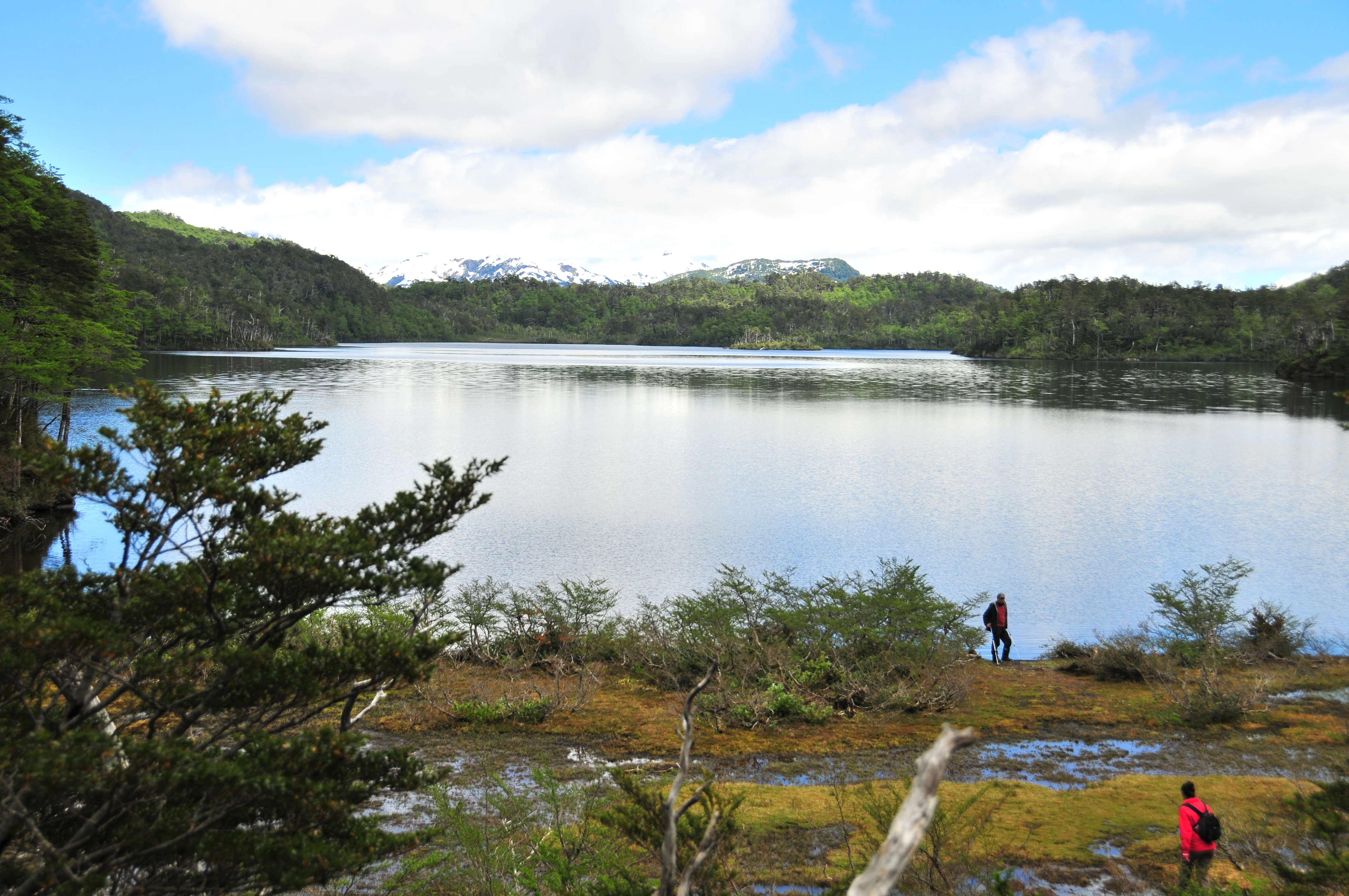A lake surrounded by forested hills with mountains in the background. Two people are standing near the water's edge.
