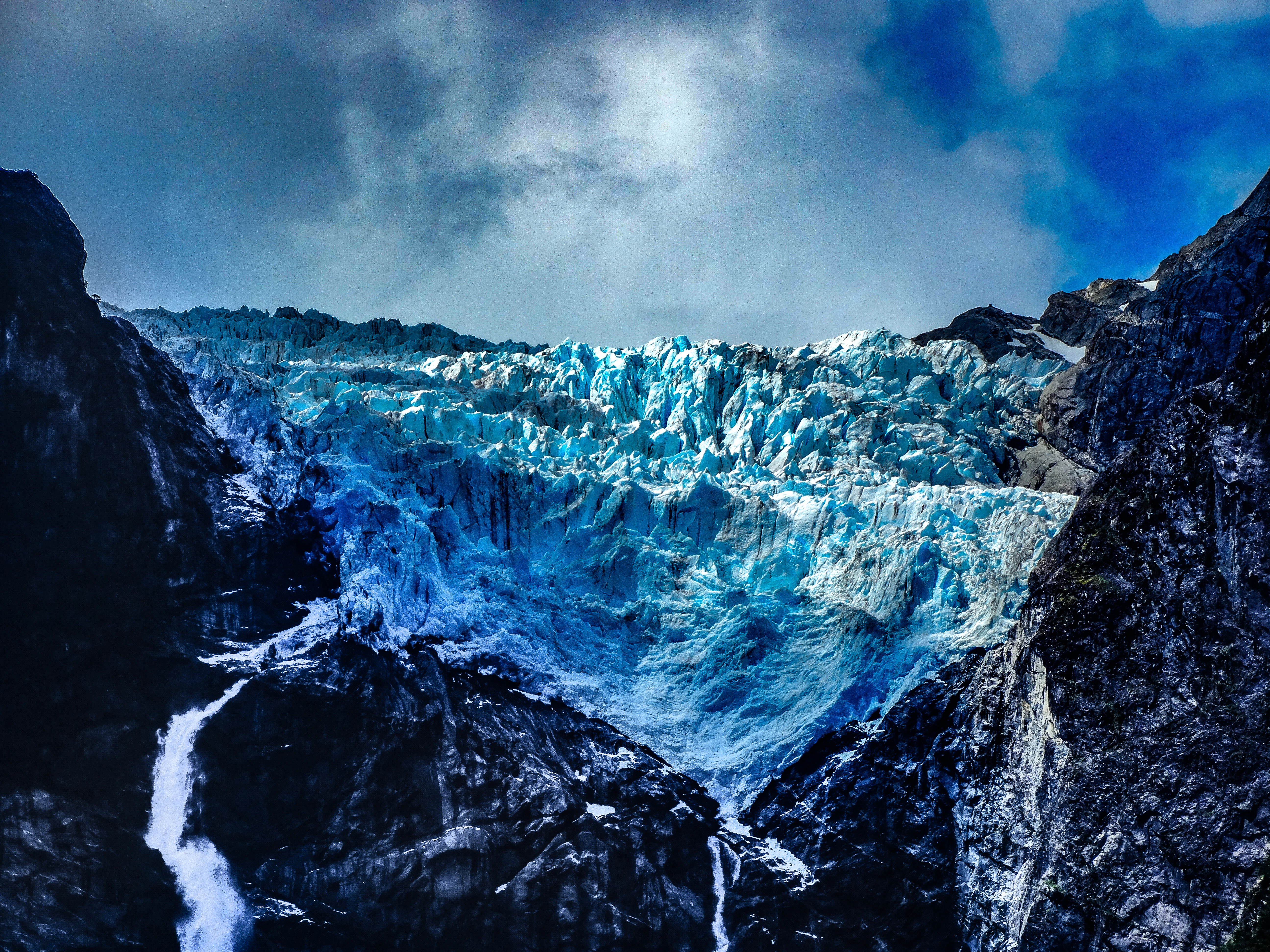 A large blue glacier cascading down between dark rocky cliffs with waterfalls flowing from its base under a partly cloudy sky