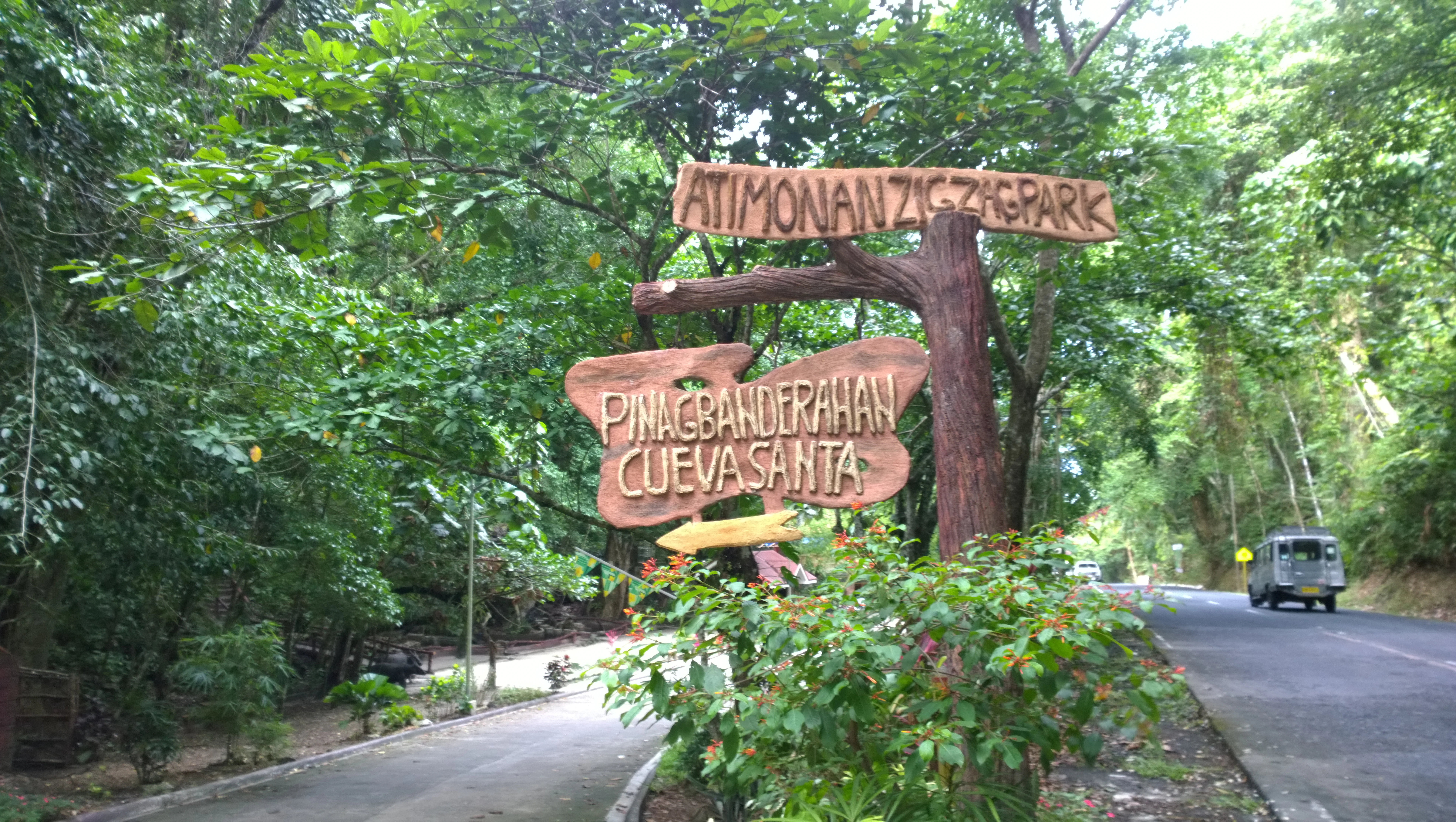 A decorative wooden sign shaped like branches and wood planks reads Atimonan Zigzag Park, Pinagbanderahan, and Cueva Santa, situated alongside a paved road in a forest setting.