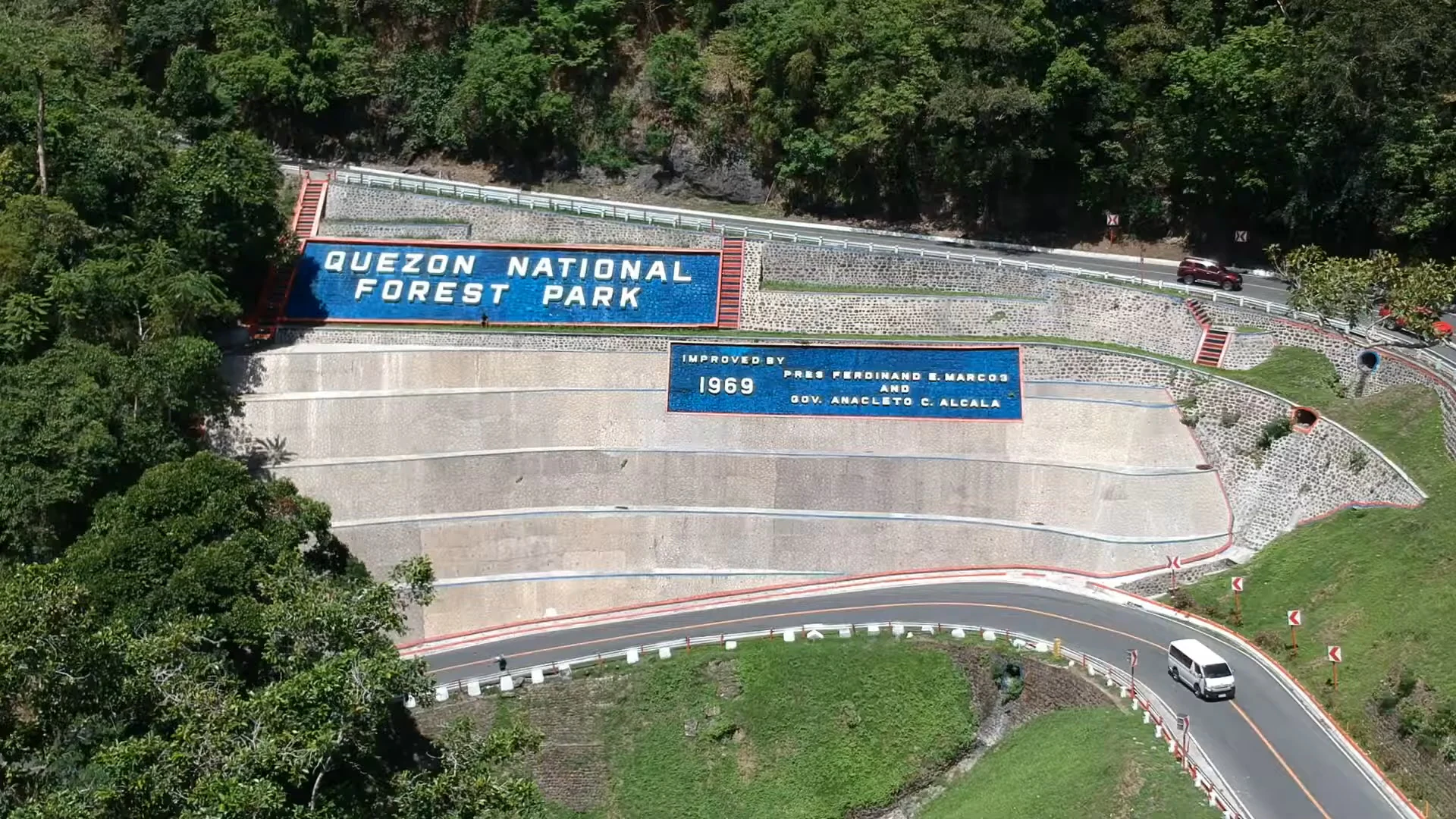 An aerial view of a curved road passing by a large concrete retaining wall featuring blue signs that read Quezon National Forest Park and indicate the year 1969.